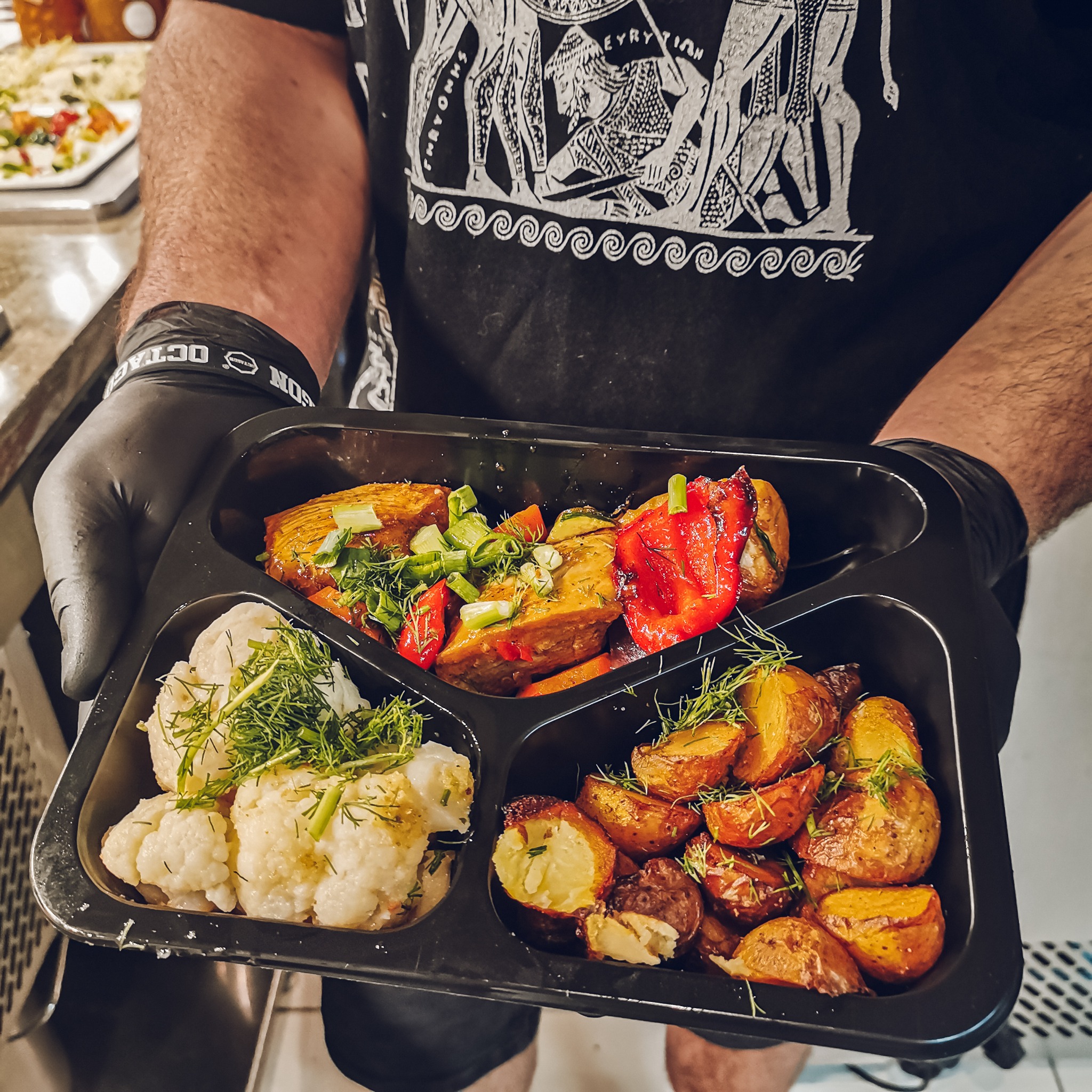 Czarny pojemnik cateringowy z czterema przegródkami: kalafior z koperkiem, pieczone ziemniaki z koperkiem, warzywa z grilla (papryka, cukinia) i kawałki mięsa posypane zielonym szczypiorkiem...