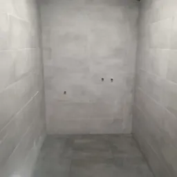 Wnętrze kabiny prysznicowej wyłożonej szarymi, matowymi płytkami ceramicznymi, z widocznymi przyłączami wodnymi na ścianie i szarą podłogą z płytek.