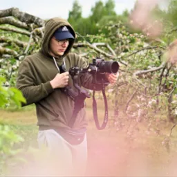 Młody mężczyzna w bluzie z kapturem i czapce z daszkiem obsługuje profesjonalną kamerę na stabilizatorze w sadzie kwitnących jabłoni.