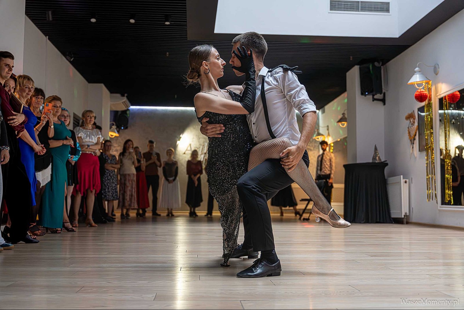 Para tańczy tango na tle publiczności w sali balowej. Kobieta w cekinowej sukni, mężczyzna w białej koszuli i czarnych spodniach. Dynamiczne ujęcie, taniec pełen ekspresji.