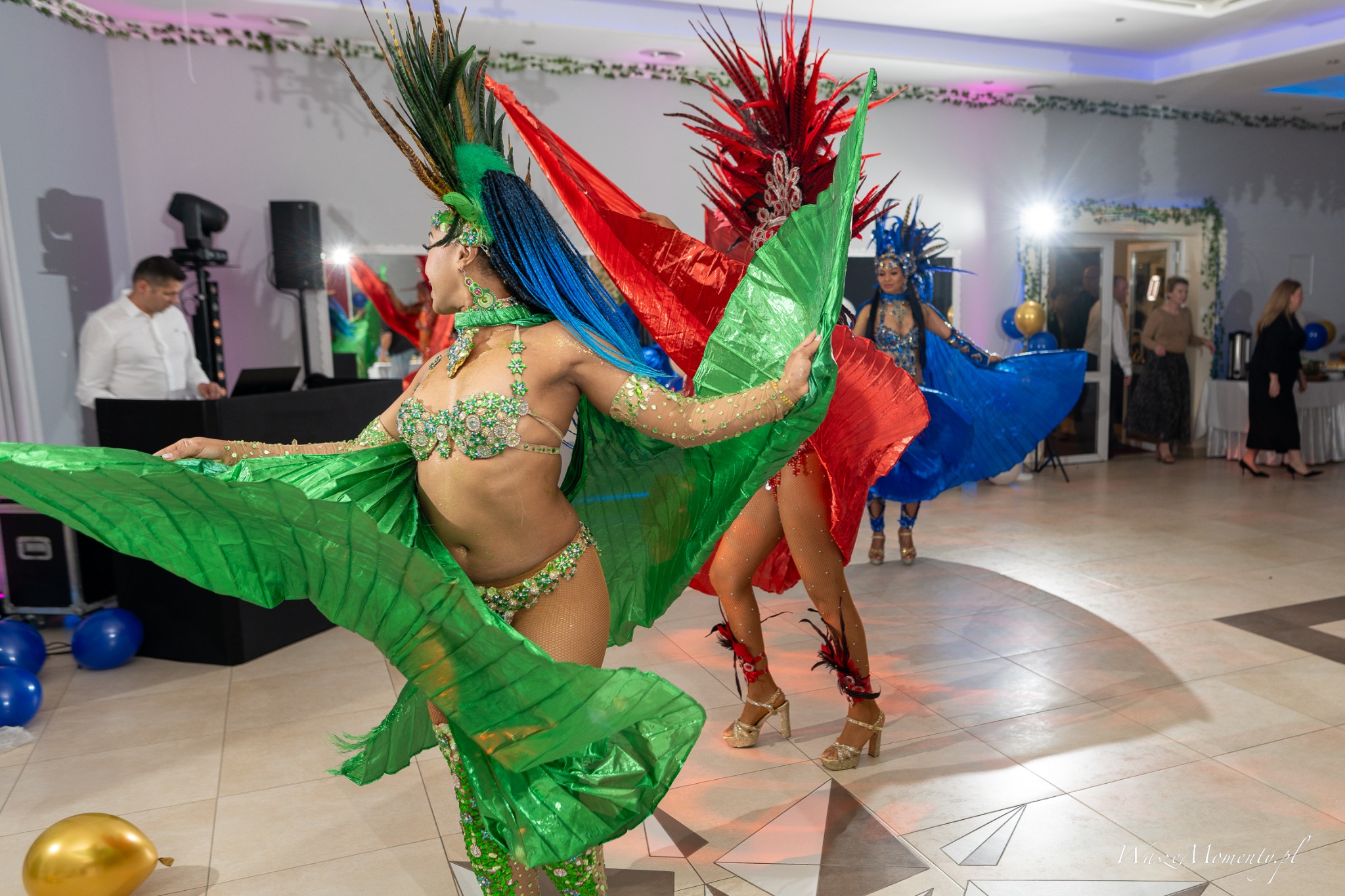Barwne tancerki w kostiumach karnawałowych prezentują układ taneczny na tle sali bankietowej z DJ-em i gośćmi. Dynamiczne ujęcie z bliska, ukazujące detale strojów.