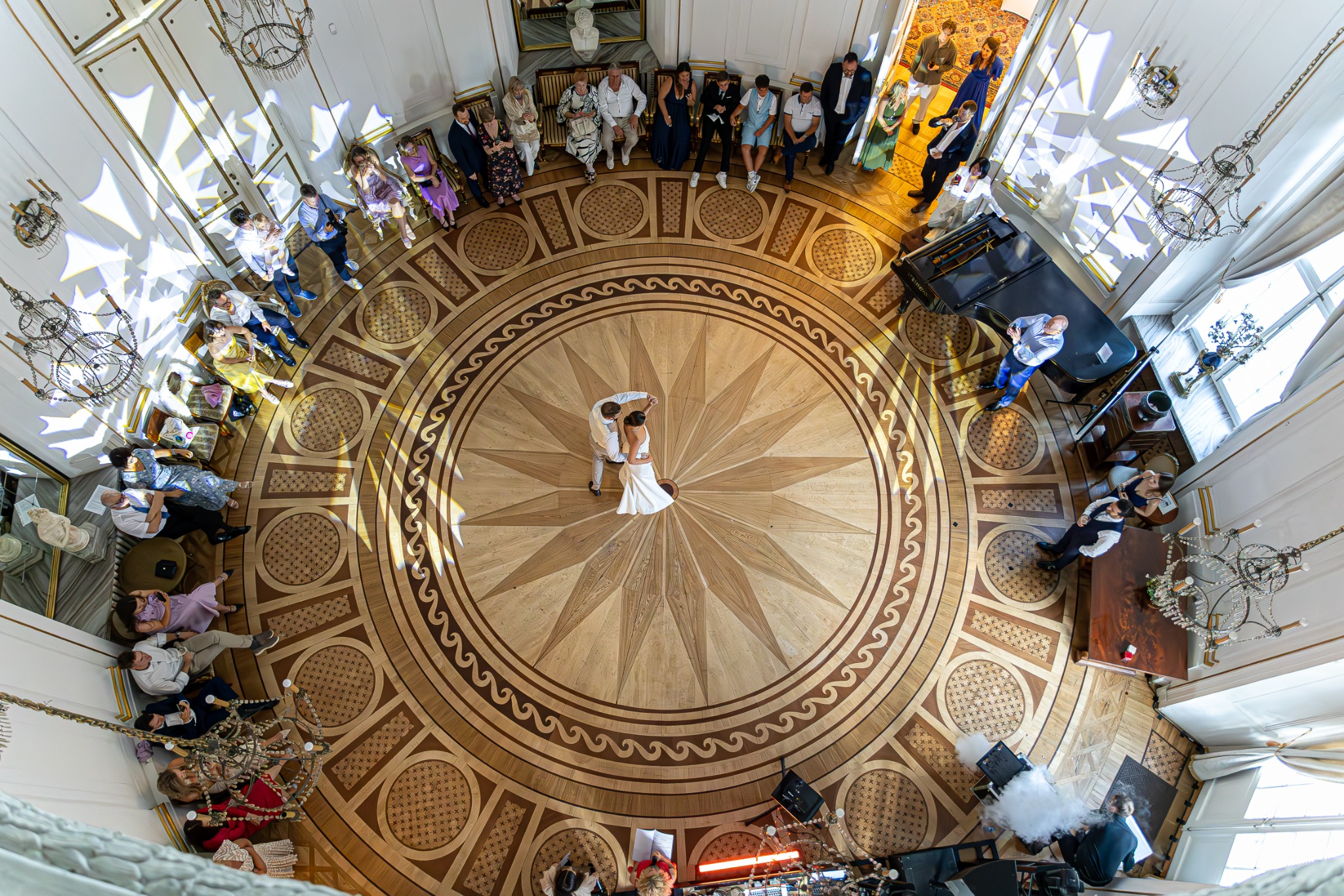 Para młoda tańczy pierwszy taniec na parkiecie z dekoracyjnym wzorem, widziana z góry. Goście siedzą wzdłuż ścian, a w tle widać pianino i oświetlenie.