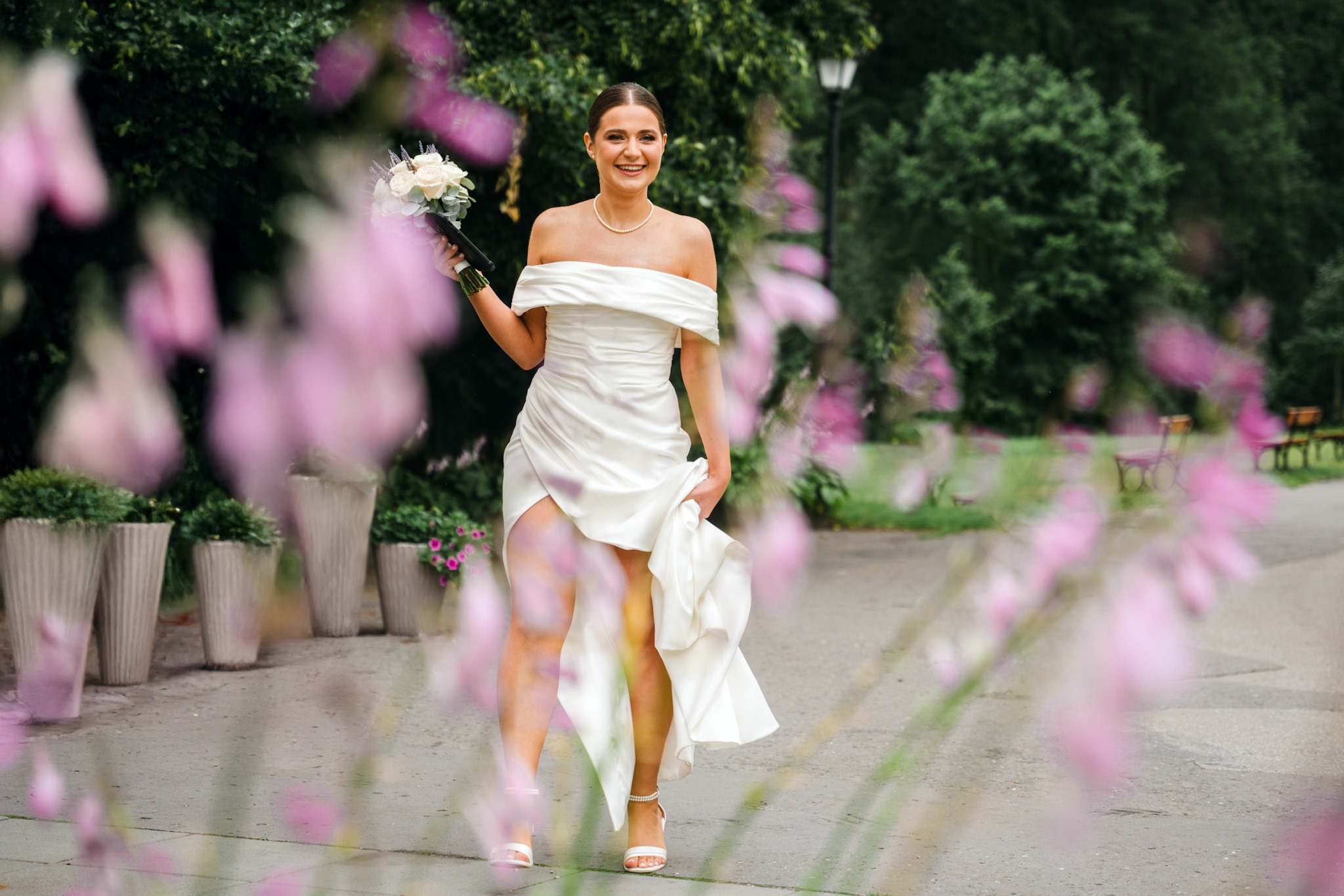 Uśmiechnięta panna młoda w białej sukni z bukietem, spacerująca po parku. Rozmyte różowe kwiaty na pierwszym planie dodają romantycznego charakteru.