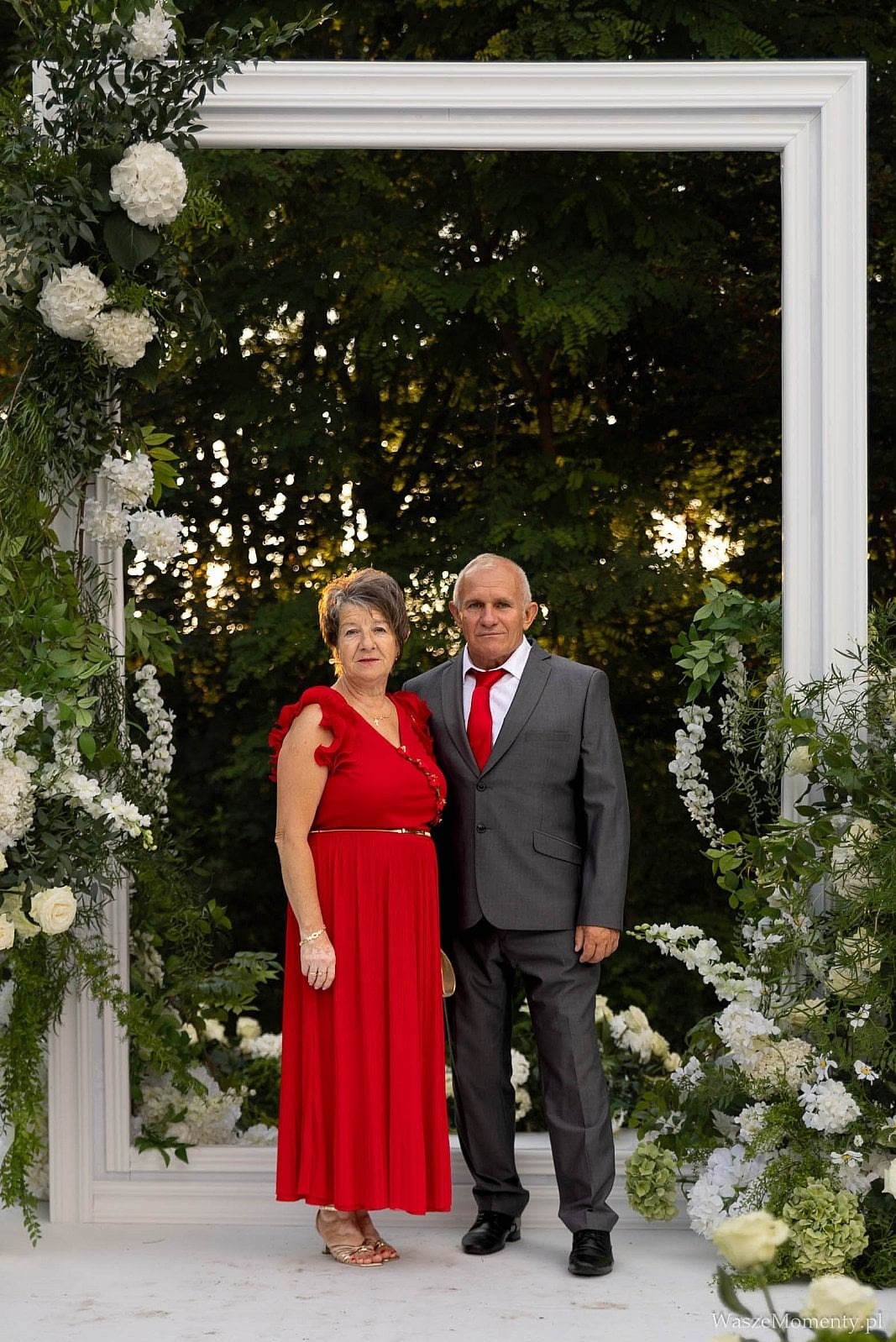 Para w średnim wieku pozuje do zdjęcia w plenerze, na tle białej ramy udekorowanej kwiatami i zielenią. Kobieta w czerwonej sukience, mężczyzna w szarym garniturze z czerwonym krawatem.