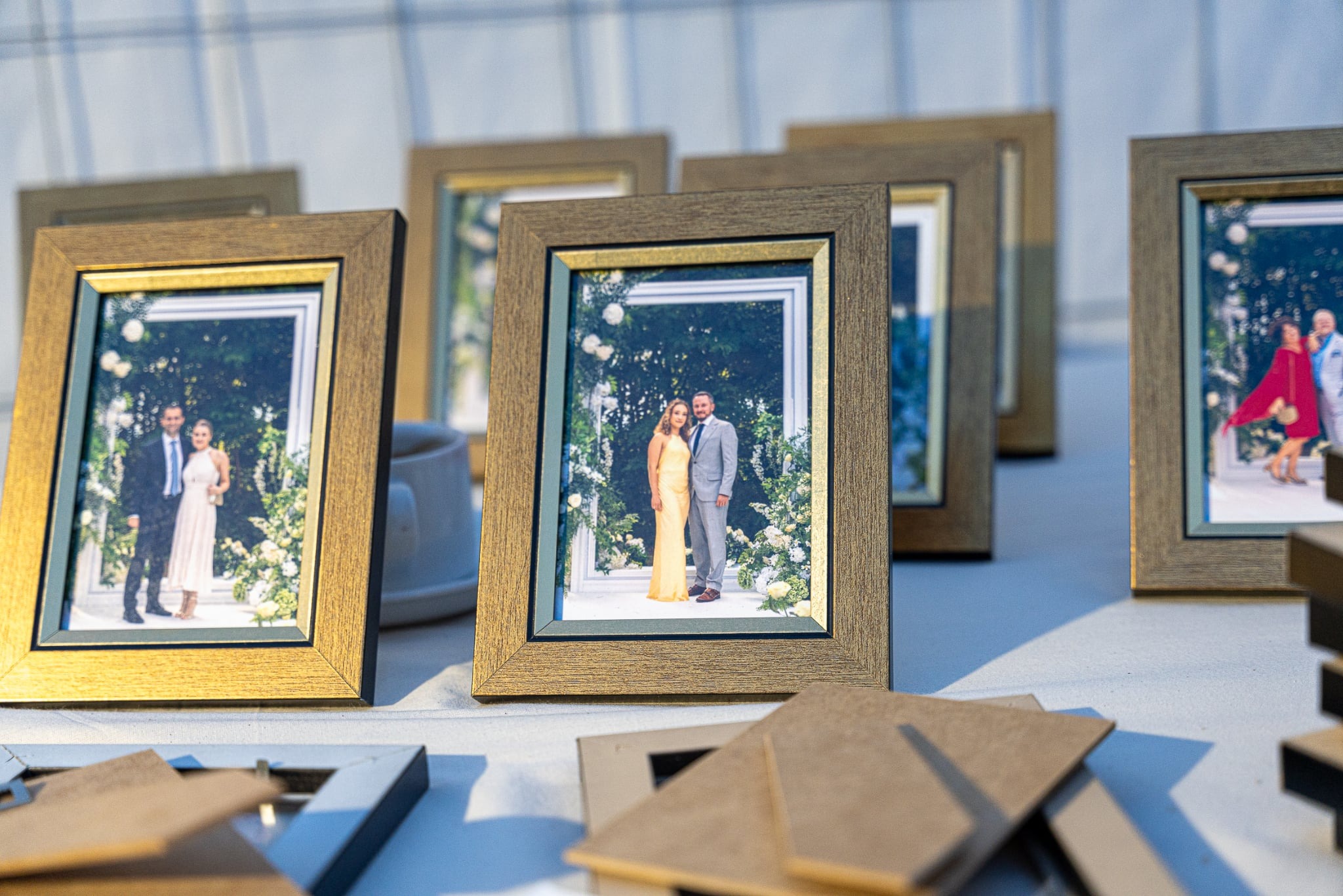 Ustawione w rzędzie oprawione zdjęcia par w strojach ślubnych, w ciepłym oświetleniu, z widocznymi elementami dekoracyjnymi i fragmentami ramek na pierwszym planie.