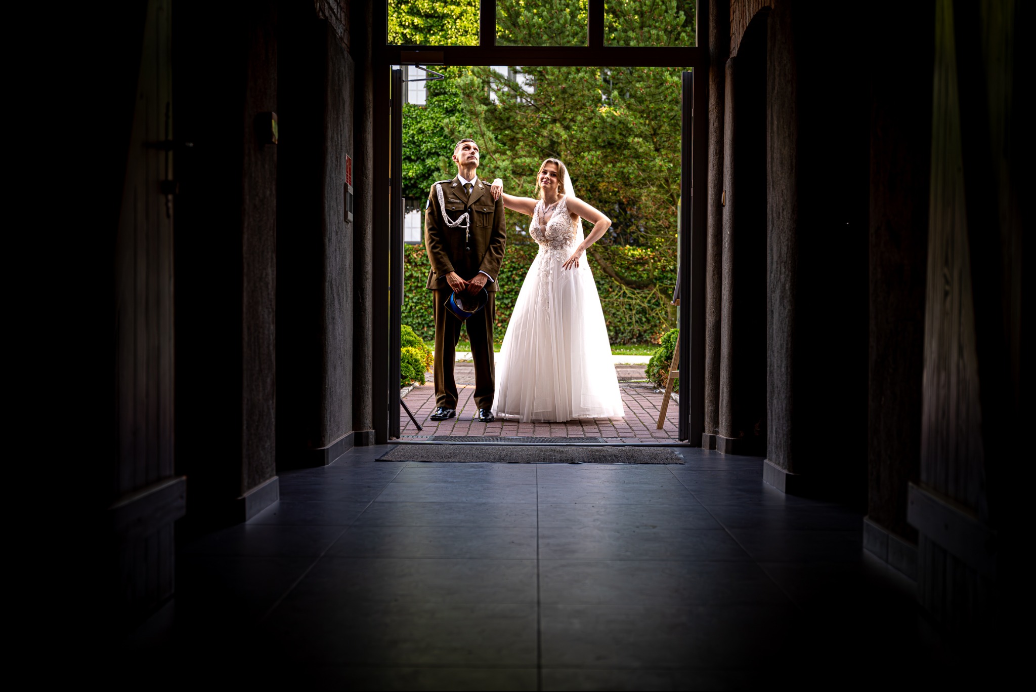 Para młoda: panna młoda w białej sukni opiera się na ramieniu pana młodego w mundurze, stoją w wejściu do budynku, patrząc w górę. Ujęcie z ciemnego wnętrza.