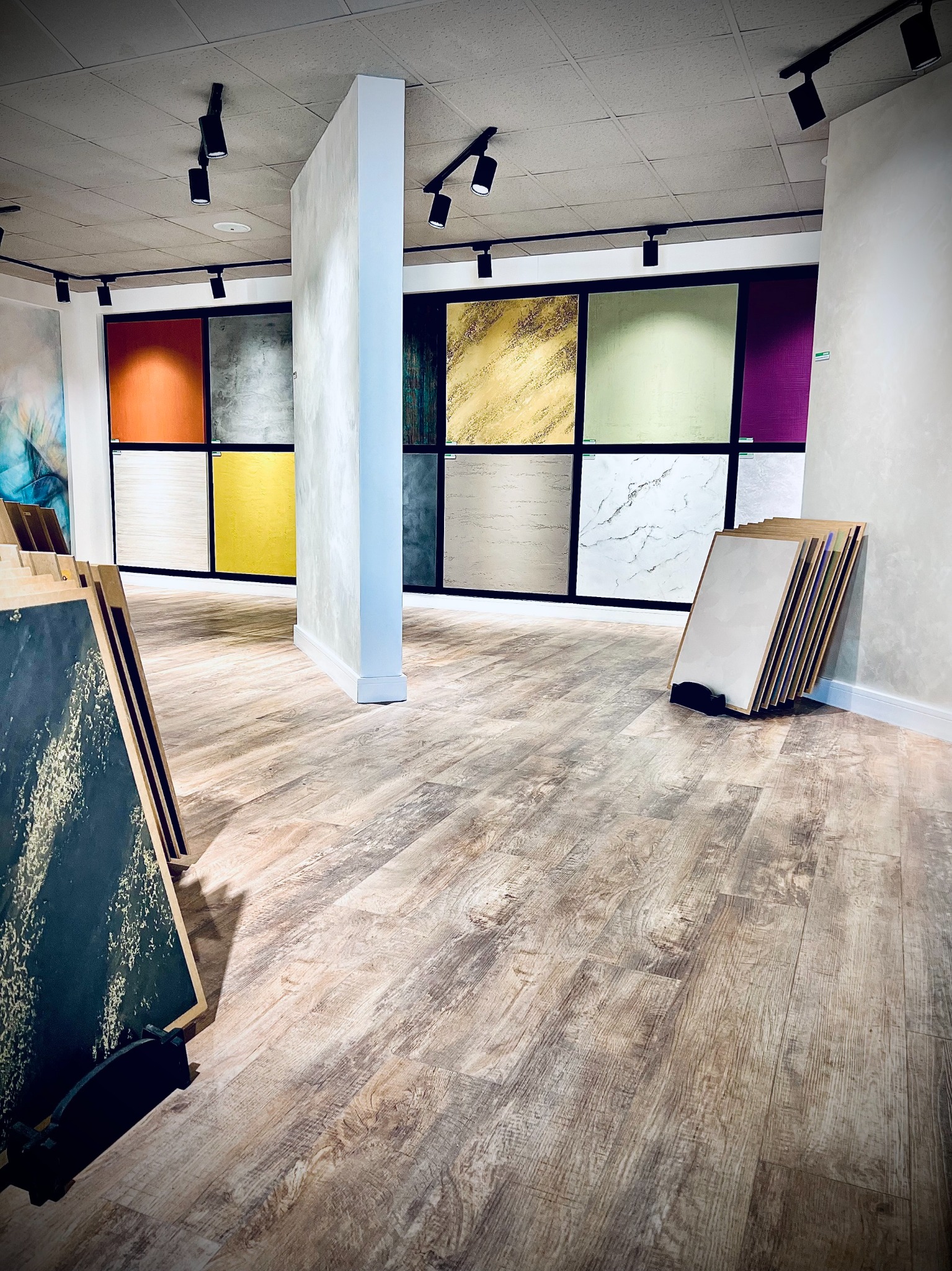 Ekspozycja próbek tynków dekoracyjnych w salonie, różne kolory i faktury, oświetlenie punktowe, podłoga imitująca drewno.