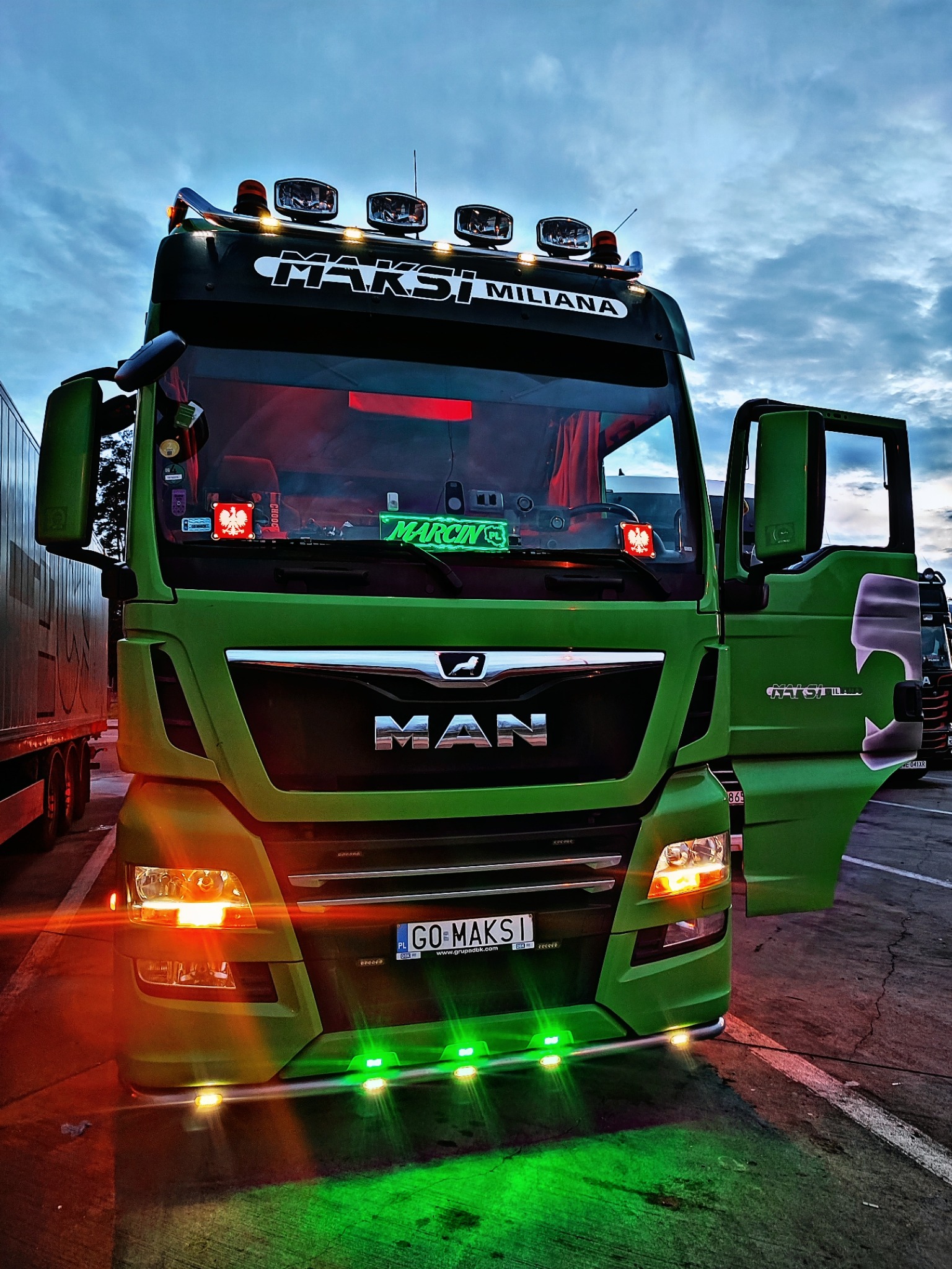 Zielony ciągnik siodłowy MAN z otwartymi drzwiami, podświetleniem LED i nazwą 'MAKSI MILIANA' na tle zachmurzonego nieba.