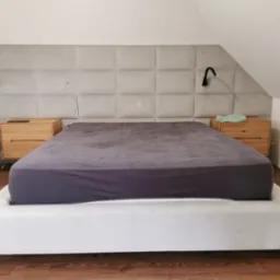 Tapicerowane wezgłowie łóżka z kwadratowych paneli w kolorze szarym, z drewnianymi szafkami nocnymi po bokach, czarna lampka nocna po prawej stronie, w pokoju na poddaszu.