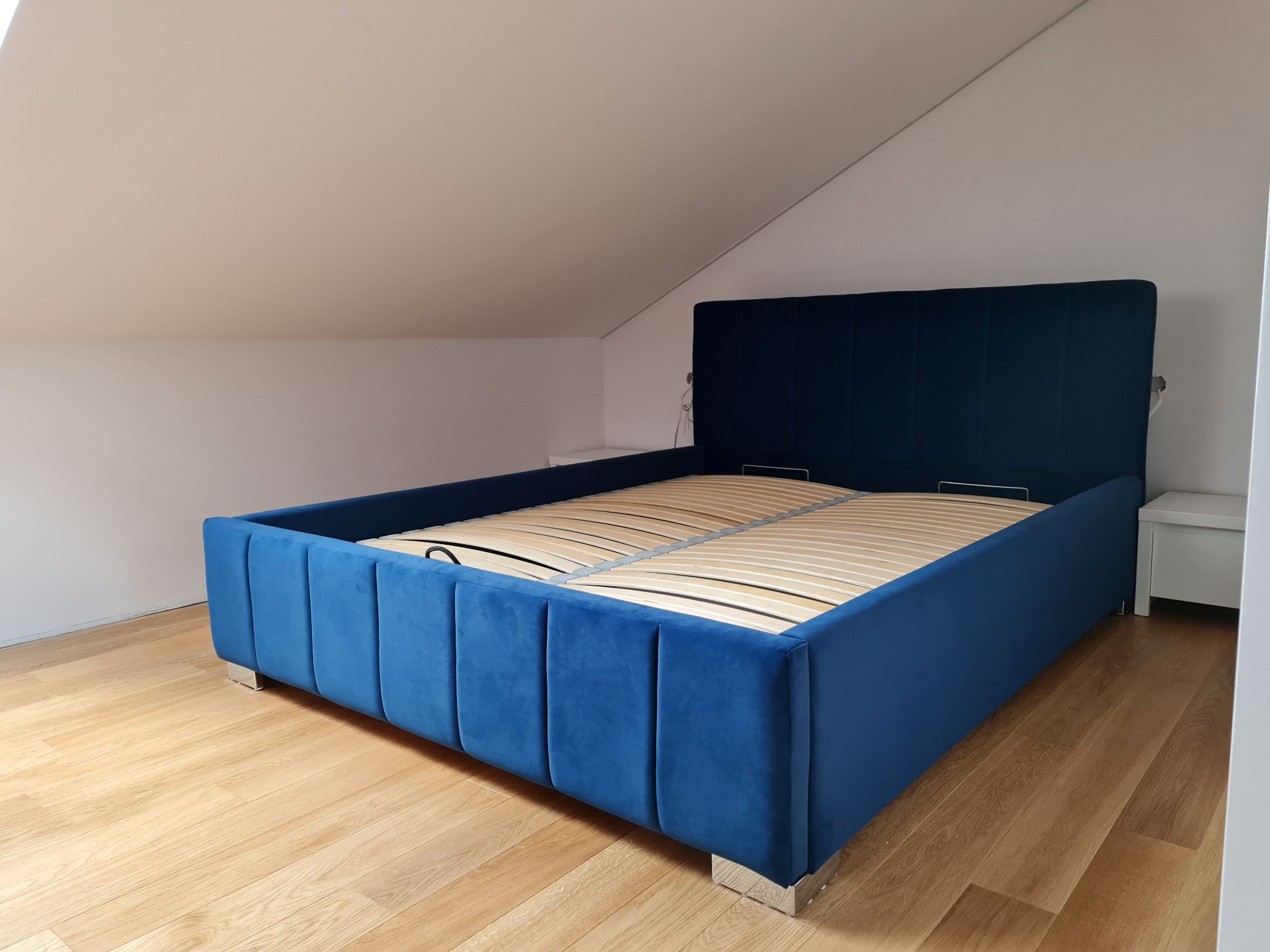 Niebieskie łóżko tapicerowane z drewnianym stelażem w pokoju na poddaszu w Łomiankach, widoczne srebrne nóżki i biała szafka nocna.