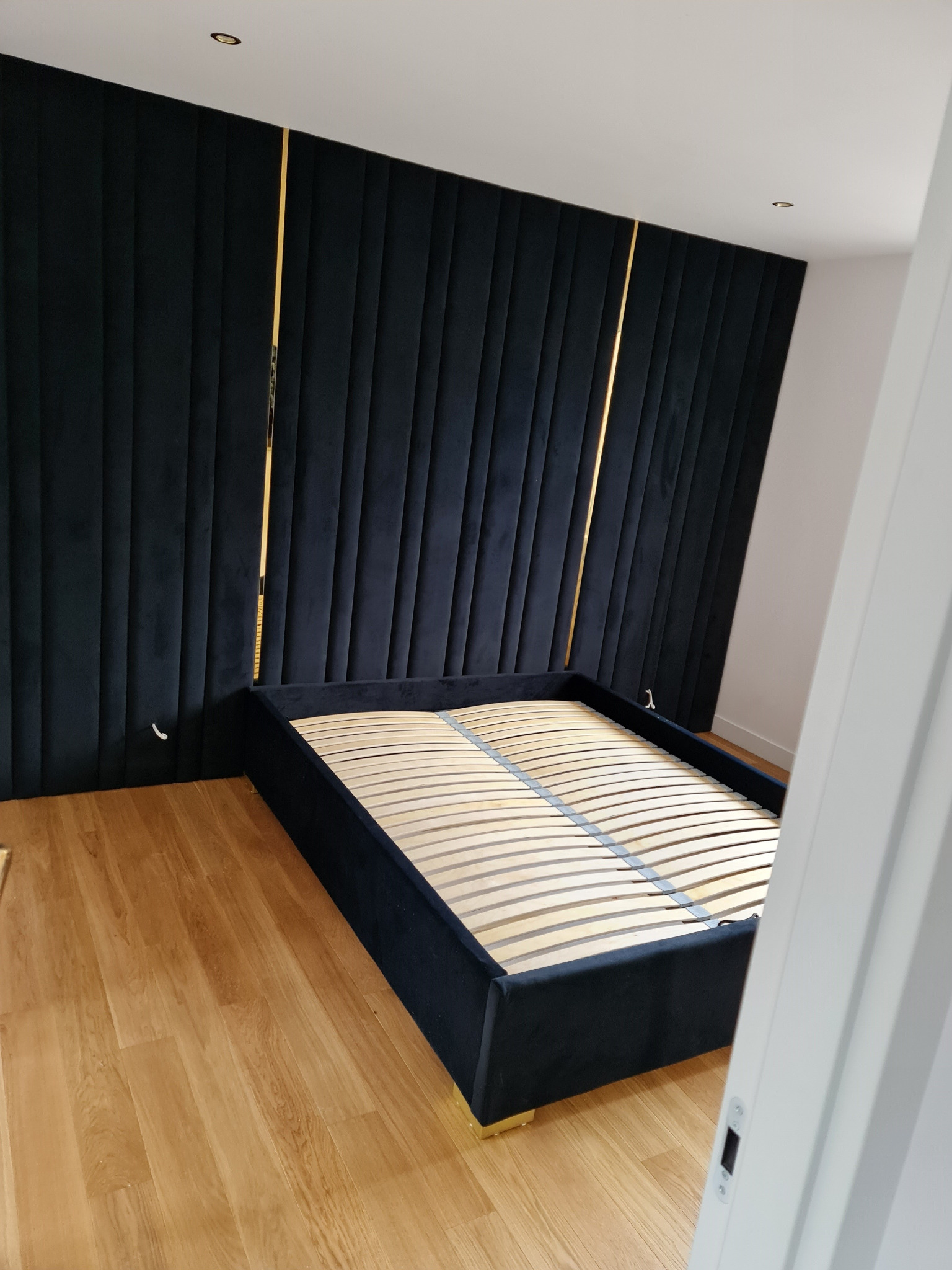 Czarne, tapicerowane łóżko z drewnianym stelażem, z wysokim zagłówkiem z pionowymi przeszyciami i złotymi akcentami, ustawione na drewnianej podłodze.