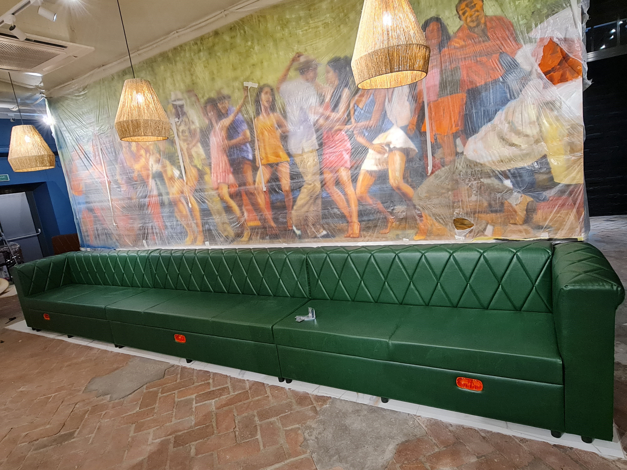 Długi, zielony, pikowany narożnik tapicerowany skórą, z widocznymi pomarańczowymi elementami oświetleniowymi w dolnej części, ustawiony na tle ściany z kolorowym muralem zakrytym folią ochronną.