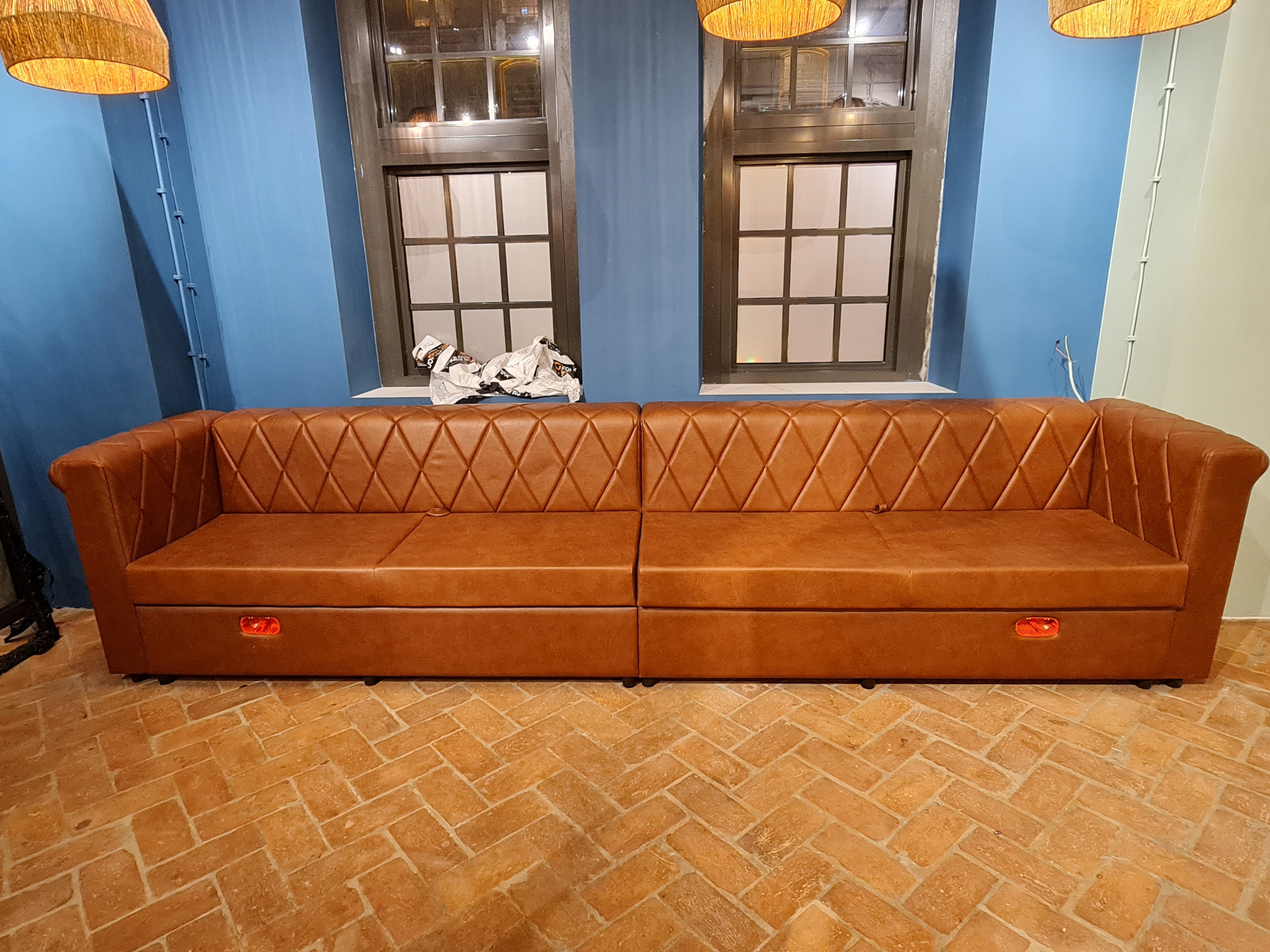 Długa, brązowa kanapa ze skóry z pikowanym oparciem w romby, umieszczona na ceglanej podłodze w pomieszczeniu z niebieskimi ścianami i dwoma oknami.