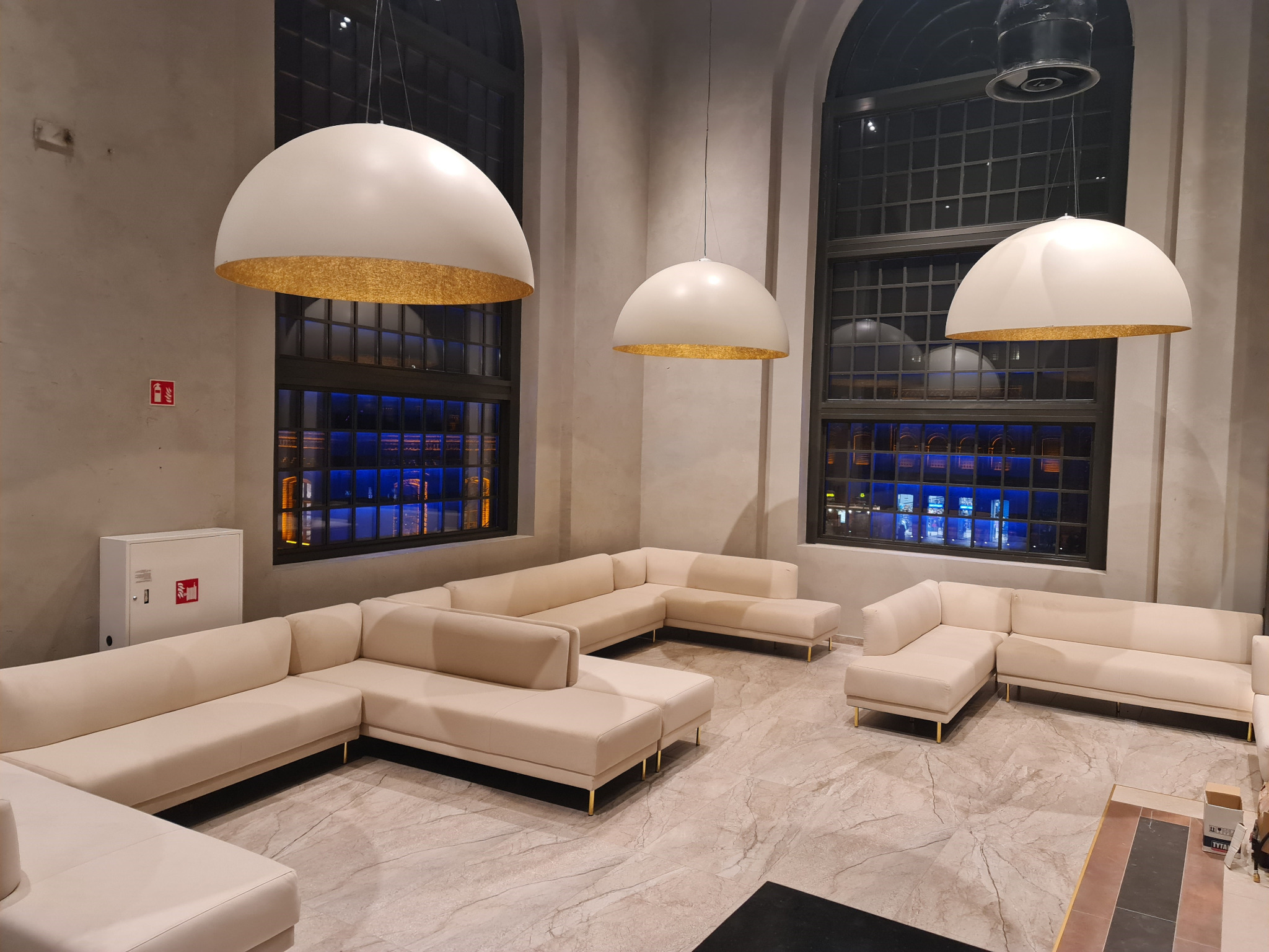 Beżowe, modułowe sofy z metalowymi nóżkami w przestronnym holu z wysokimi oknami i dużymi, kulistymi lampami z elementami w kolorze złota.