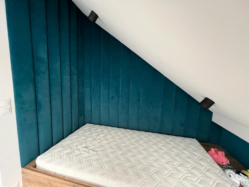 Zagłówek z paneli tapicerowanych w kolorze butelkowej zieleni, dopasowany do skosu dachu, z materacem i zabawkami w tle.