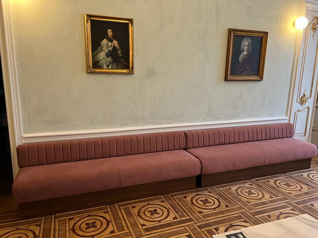 Długi, różowy, welurowy siedzisko z pionowymi przeszyciami, wkomponowane w ścianę, pod dwoma portretami w złotych ramach, na tle wzorzystej drewnianej podłogi.