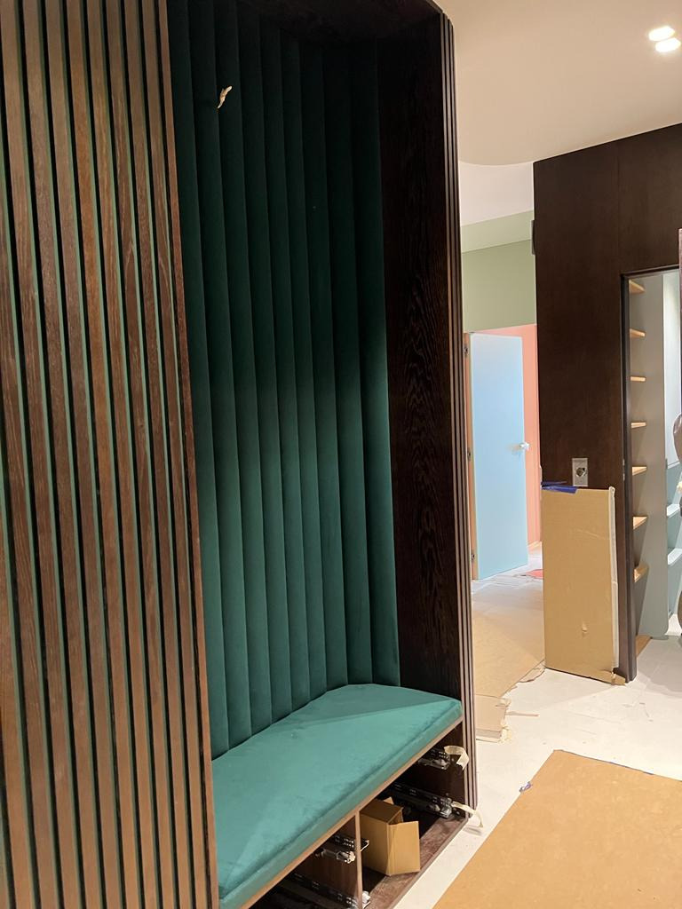 Wnęka z siedziskiem obita zielonym welurem, otoczona drewnianymi lamelami w kolorze ciemnego orzecha, widok na korytarz w trakcie wykańczania.