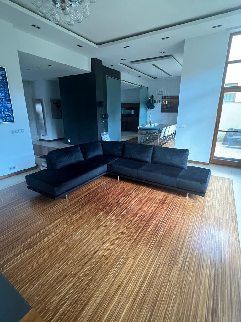 Czarna, welurowa sofa narożna z niskim oparciem i metalowymi nóżkami w przestronnym salonie z drewnianą podłogą i dużymi oknami.