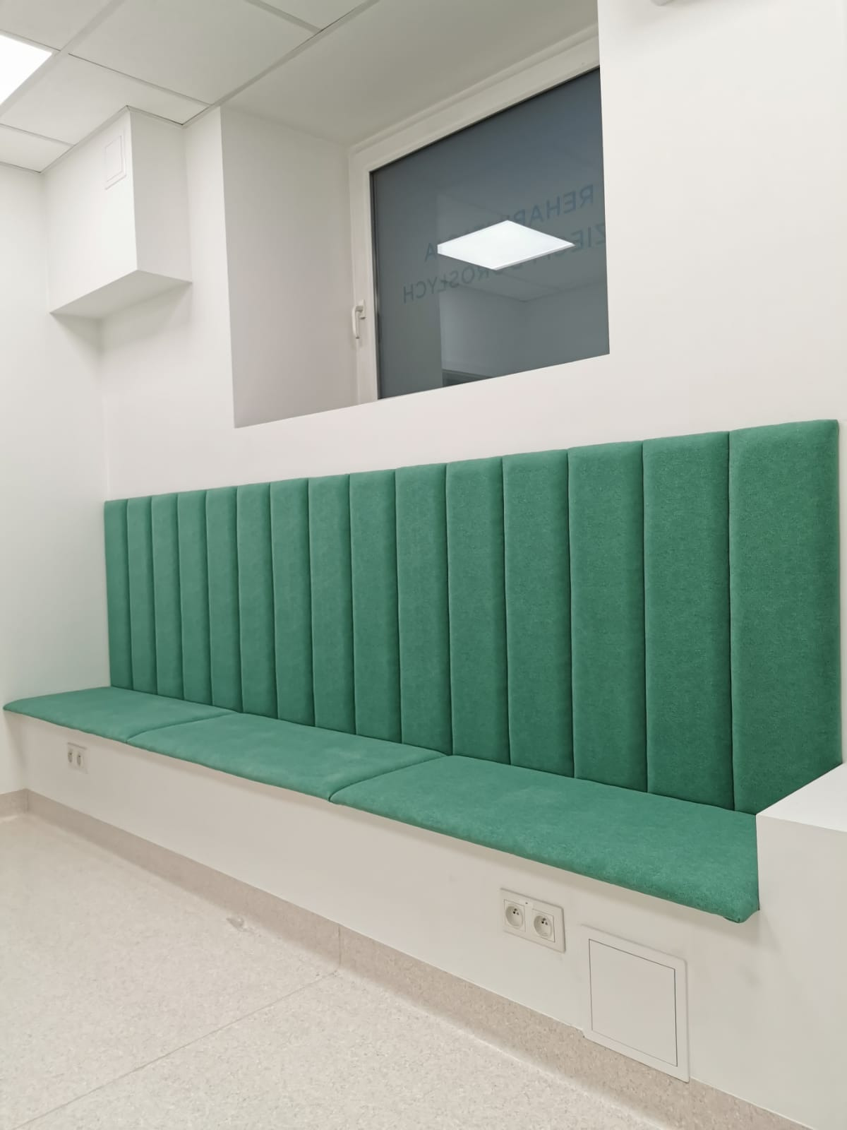 Długa, zielona ławka tapicerowana w poczekalni, z wysokim oparciem podzielonym na pionowe panele, zamontowana wzdłuż ściany z oknem.