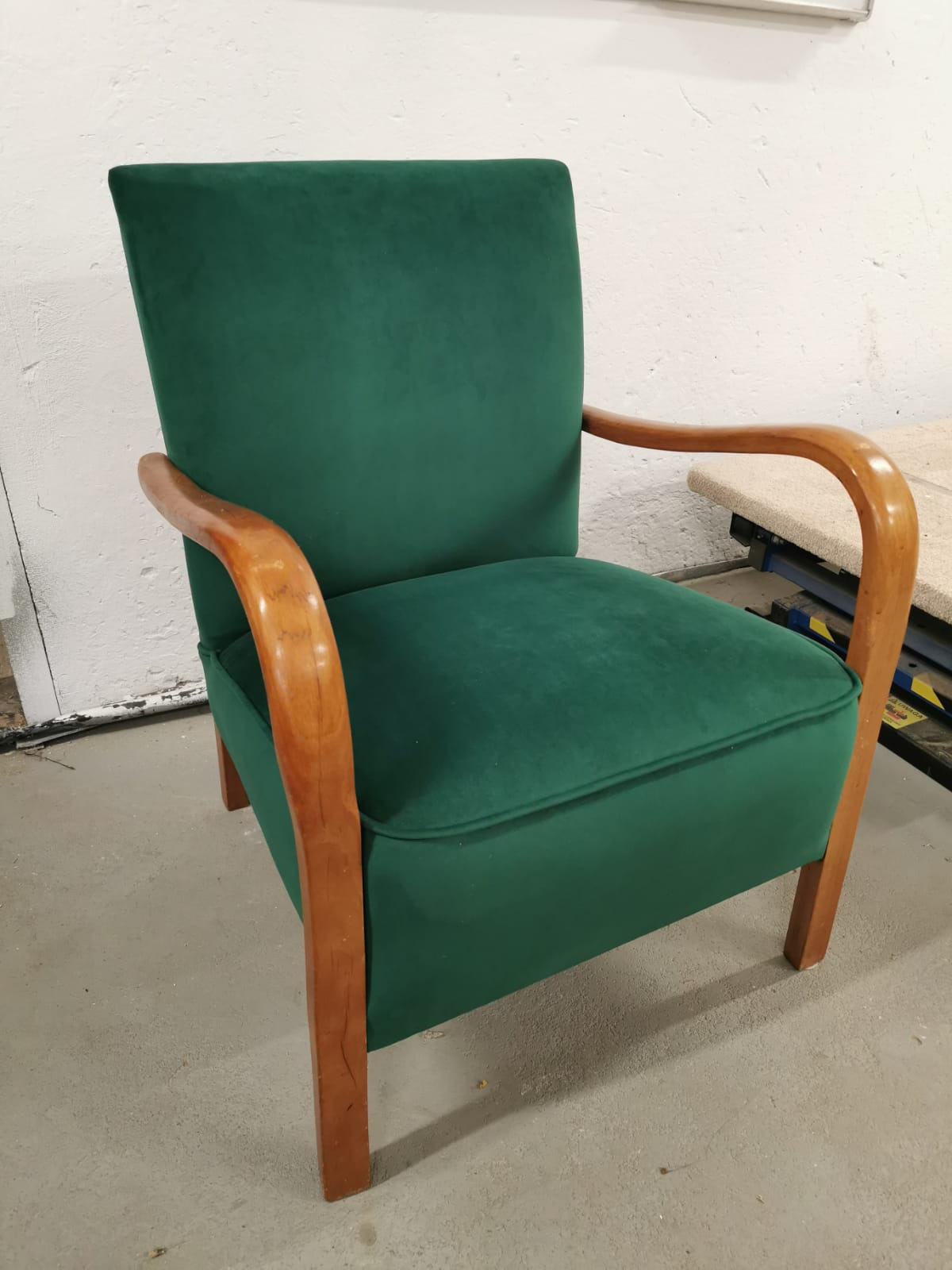 Zielony fotel tapicerowany z drewnianymi podłokietnikami w stylu retro, stojący na szarym betonowym podłożu.