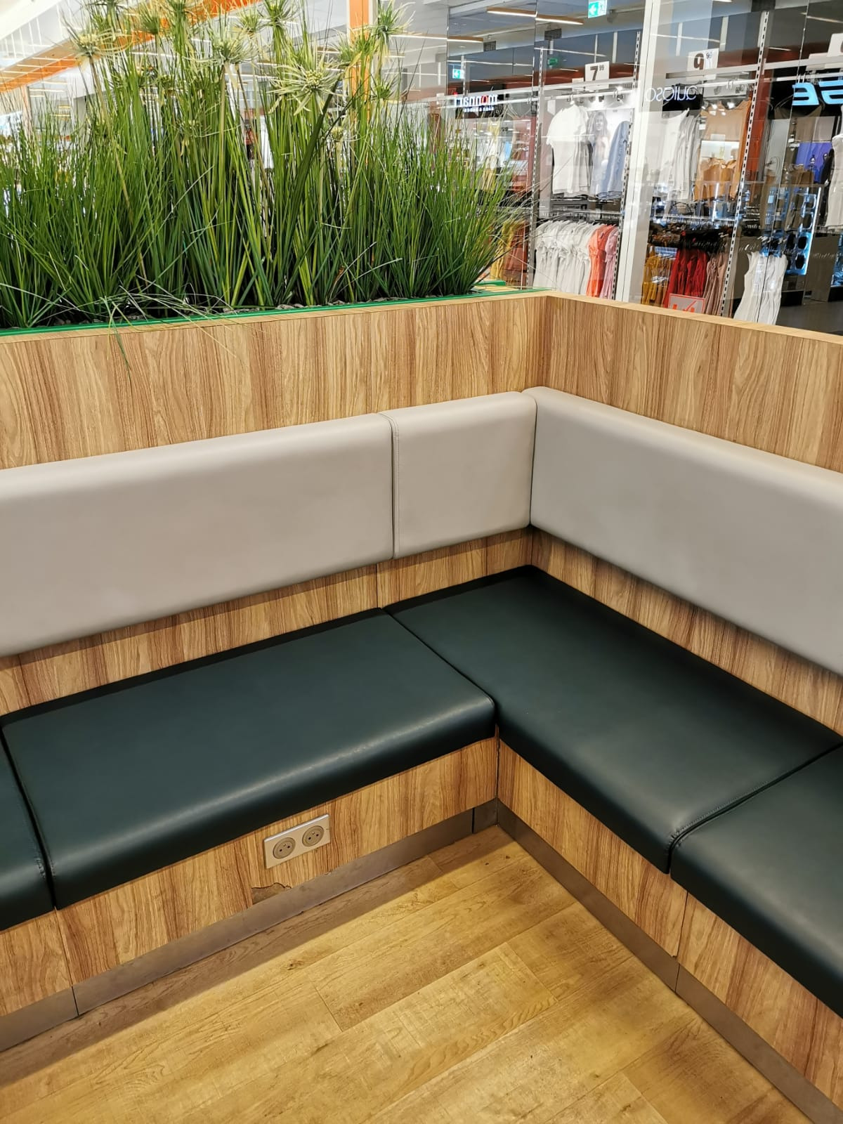 Wnętrze sklepu z narożną, tapicerowaną ławką w kolorze butelkowej zieleni i szarą ścianką, obudowaną drewnopodobną okładziną, z wbudowanymi gniazdkami elektrycznymi i donicą z wysokimi trawami.