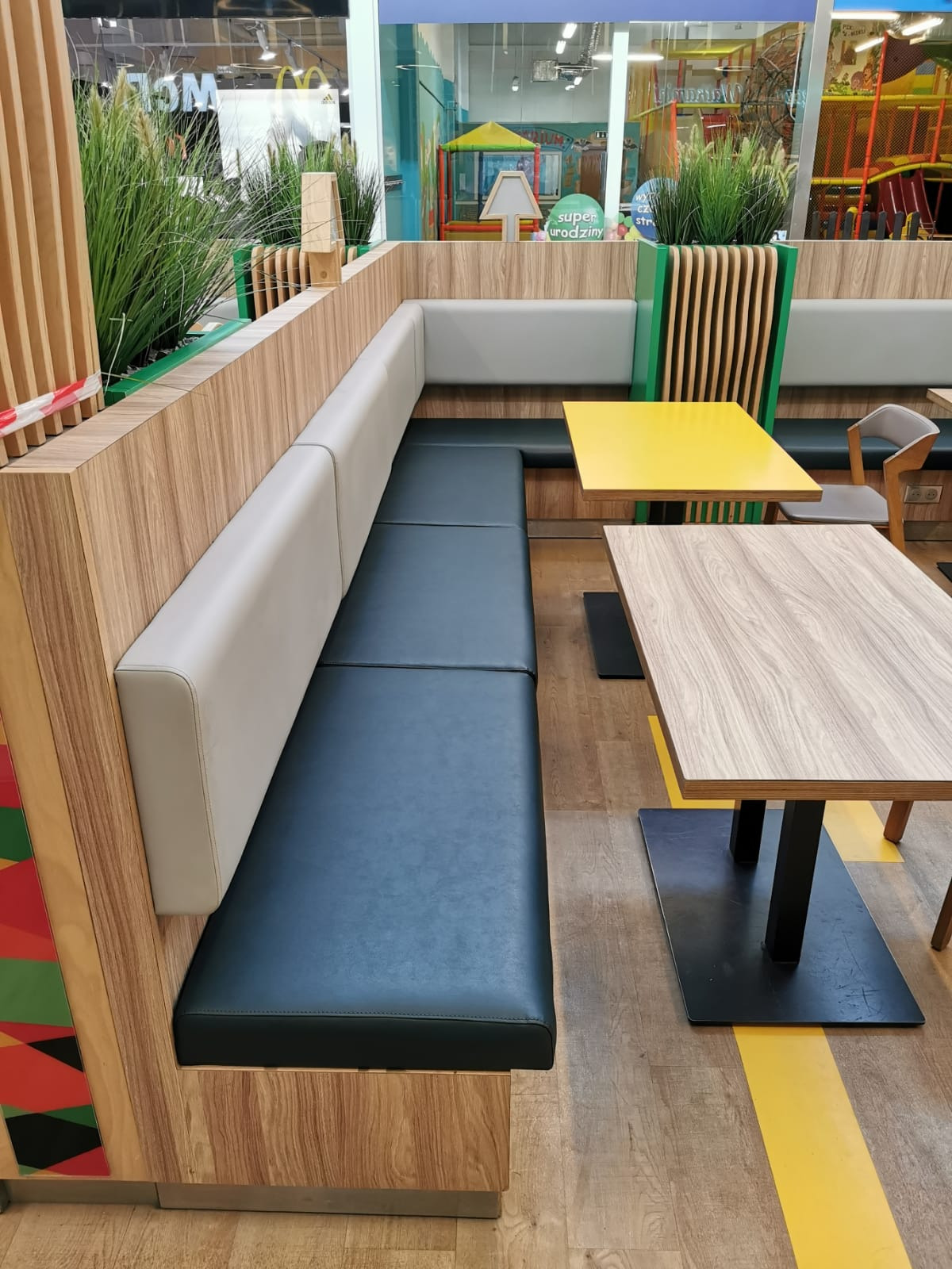 Wnętrze restauracji z długą, tapicerowaną ławą w kolorze granatowym, wspartą na drewnianej konstrukcji, obok stoliki z żółtym i drewnopodobnym blatem, a w tle plac zabaw.
