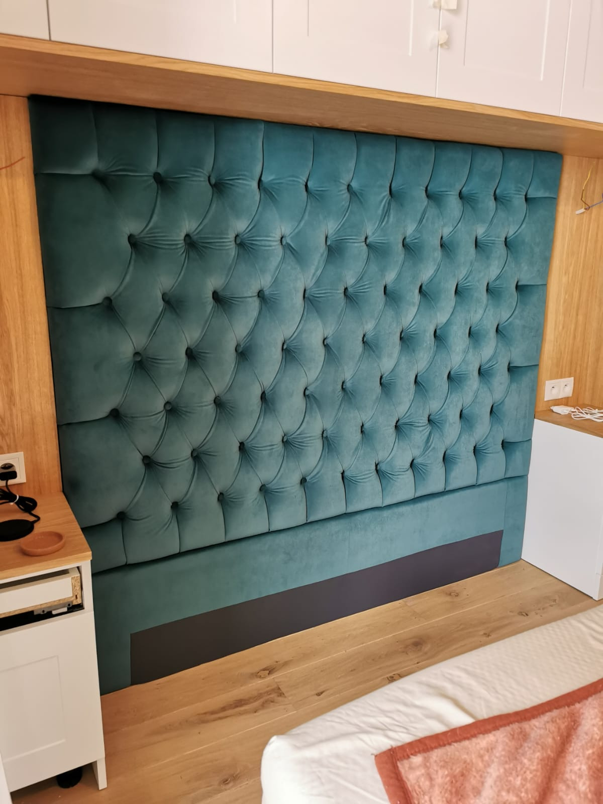 Turkusowy, tapicerowany zagłówek z pikowaniem w stylu Chesterfield, zamontowany na drewnianej ramie, widoczny fragment szafki nocnej i łóżka.