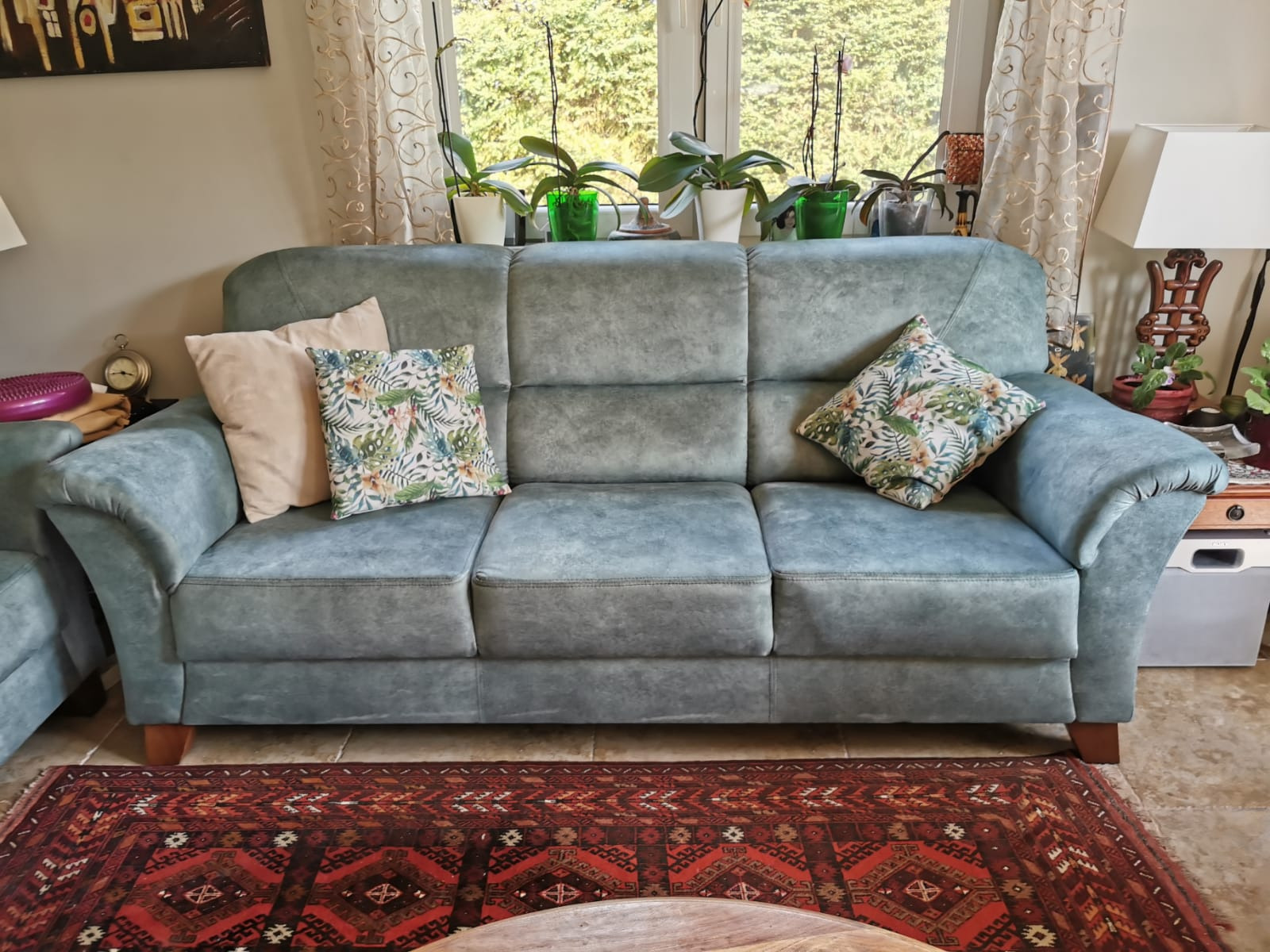 Trzyosobowa sofa tapicerowana w odcieniach szarości z poduszkami w kwiatowe wzory, ustawiona na orientalnym dywanie, z roślinami doniczkowymi na parapecie okna w tle.