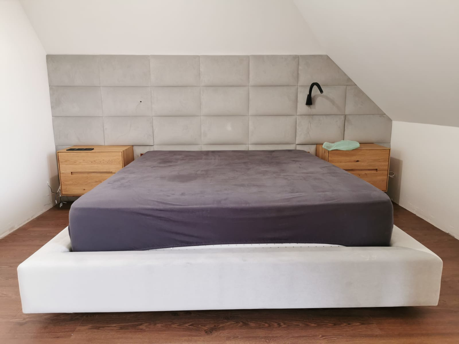 Tapicerowane wezgłowie łóżka z kwadratowych paneli w kolorze szarym, z drewnianymi szafkami nocnymi po bokach, czarna lampka nocna po prawej stronie, w pokoju na poddaszu.