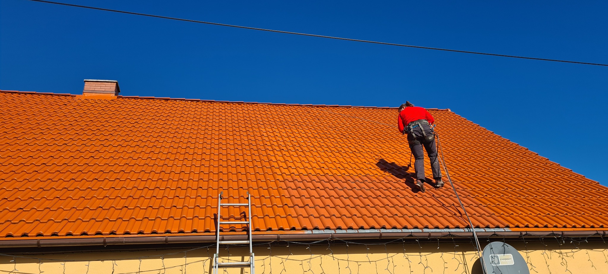 Pracownik w czerwonej koszuli, zabezpieczony linami, maluje pomarańczowy dach z dachówki ceramicznej na tle błękitnego nieba. Widoczna drabina oparta o rynnę.
