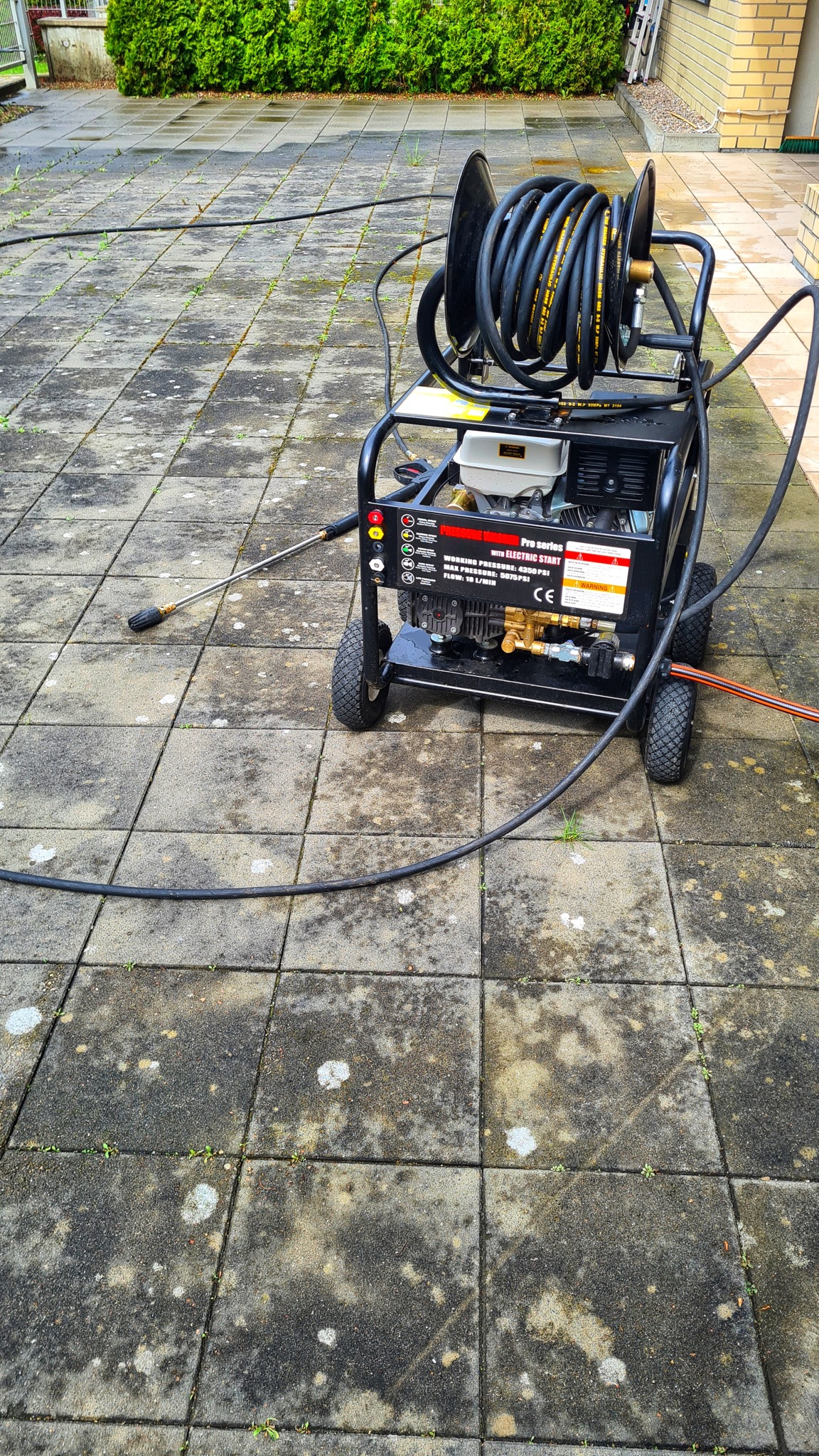 Profesjonalna myjka ciśnieniowa z nawiniętym wężem, gotowa do użycia na kostce brukowej z widocznymi zabrudzeniami i mchem w fugach.