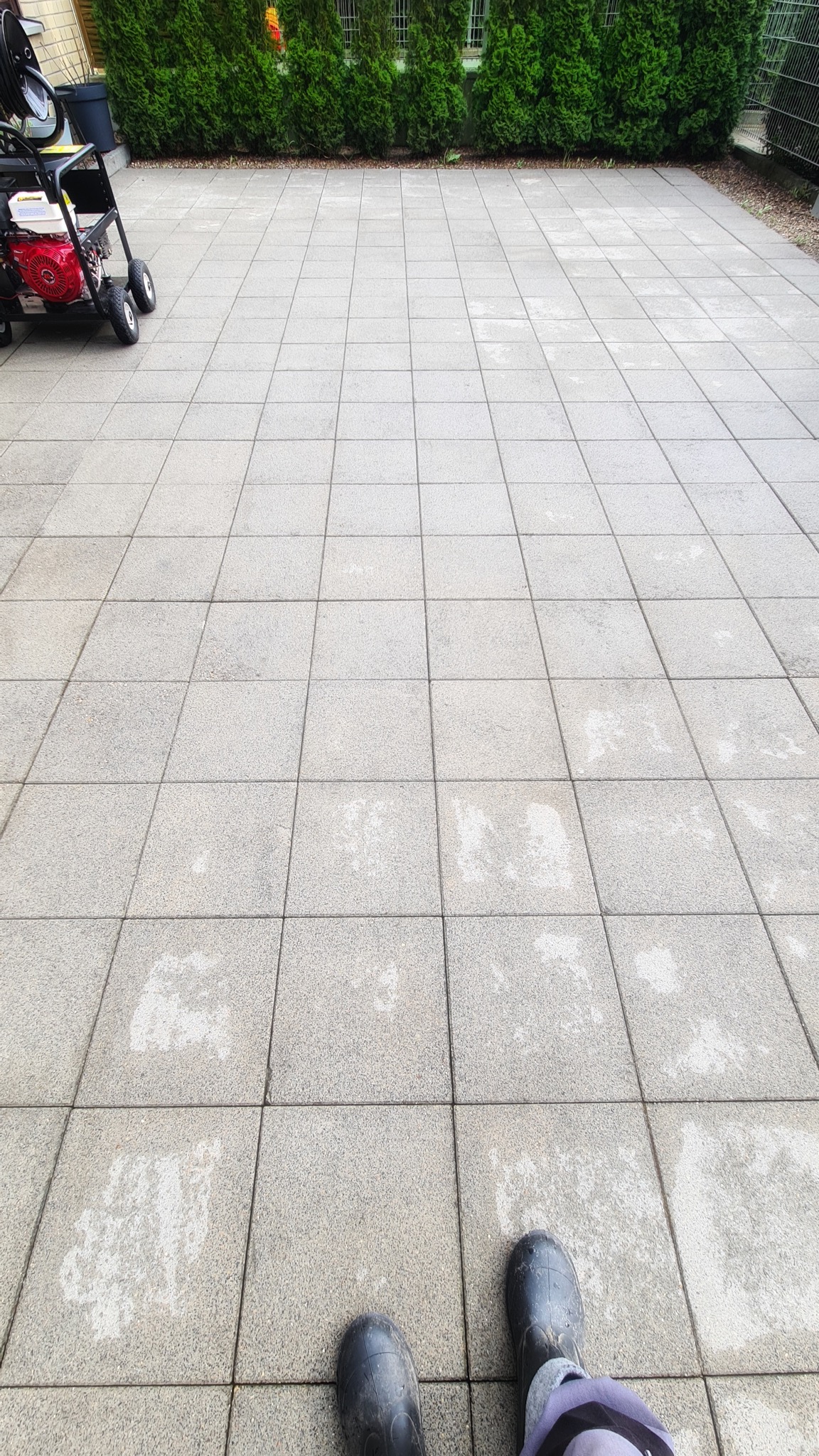 Szara kostka brukowa częściowo umyta, z widocznymi śladami po czyszczeniu, maszyna ciśnieniowa w tle i stopy w czarnych gumowcach na dole kadru.