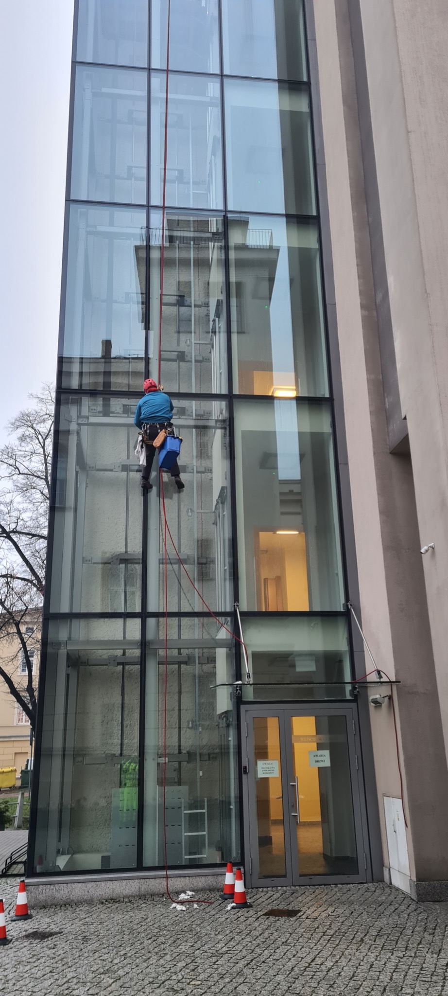 Pracownik na linie zabezpieczającej, w kasku i niebieskiej kurtce, czyści szklaną fasadę budynku. Widoczne pachołki ostrzegawcze i wejście do budynku.