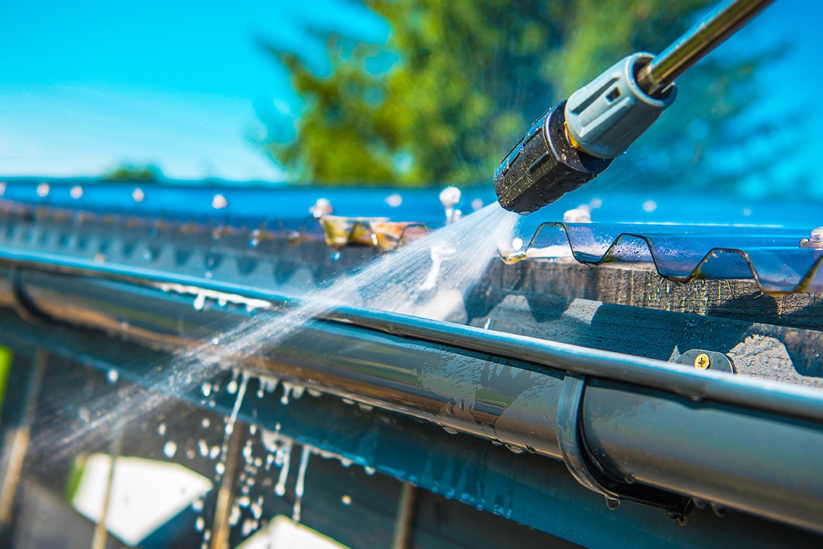 Dysza myjki ciśnieniowej kieruje strumień wody na zabrudzone rynny dachowe z widocznymi elementami mocującymi, na tle błękitnego nieba i zieleni drzew.