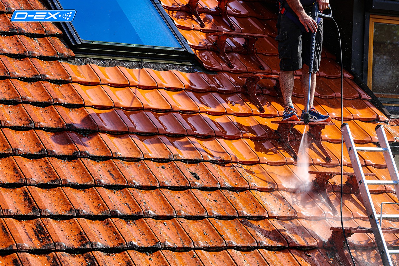Czerwona dachówka ceramiczna czyszczona myjką ciśnieniową, widoczna aluminiowa drabina, fragment okna dachowego i elewacji budynku w tle.