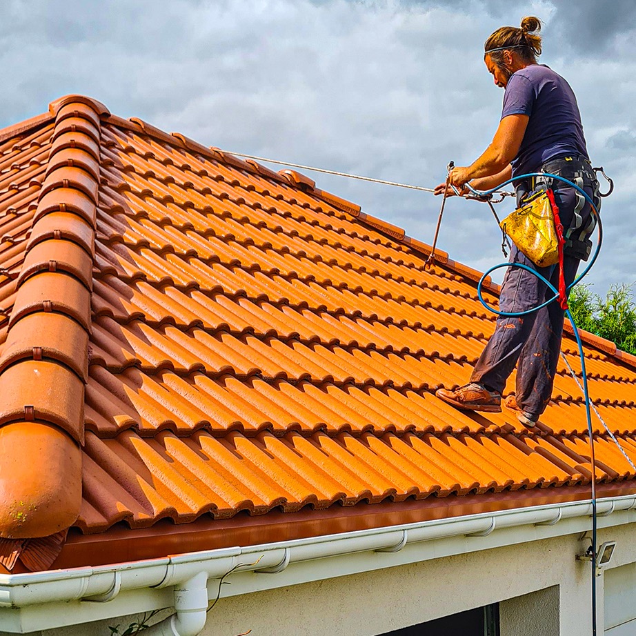 Pracownik zabezpieczony linami na pomarańczowym dachu z dachówki ceramicznej, widoczne białe rynny i fragment elewacji domu.
