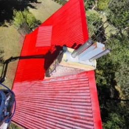 Czerwony, blaszany dach z kominem w trakcie renowacji, częściowo pomalowany, widoczne ślady starej farby i zabezpieczenia komina taśmą malarską, ujęcie z góry z widocznym otoczeniem drzew i trawnika.