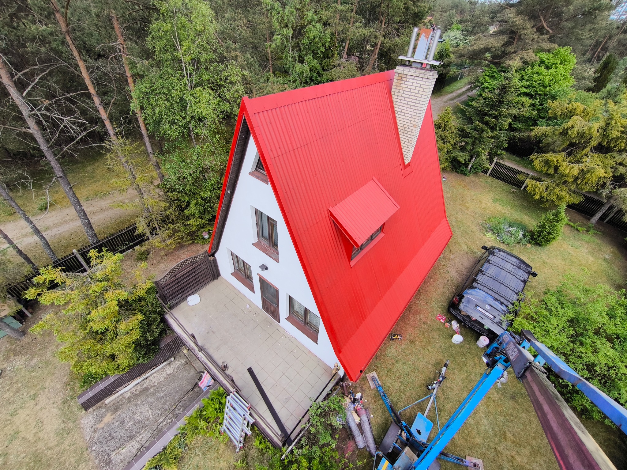 Dom z czerwonym, świeżo pomalowanym dachem, widok z góry z podnośnika koszowego, z widocznym samochodem i narzędziami pracy na trawniku.