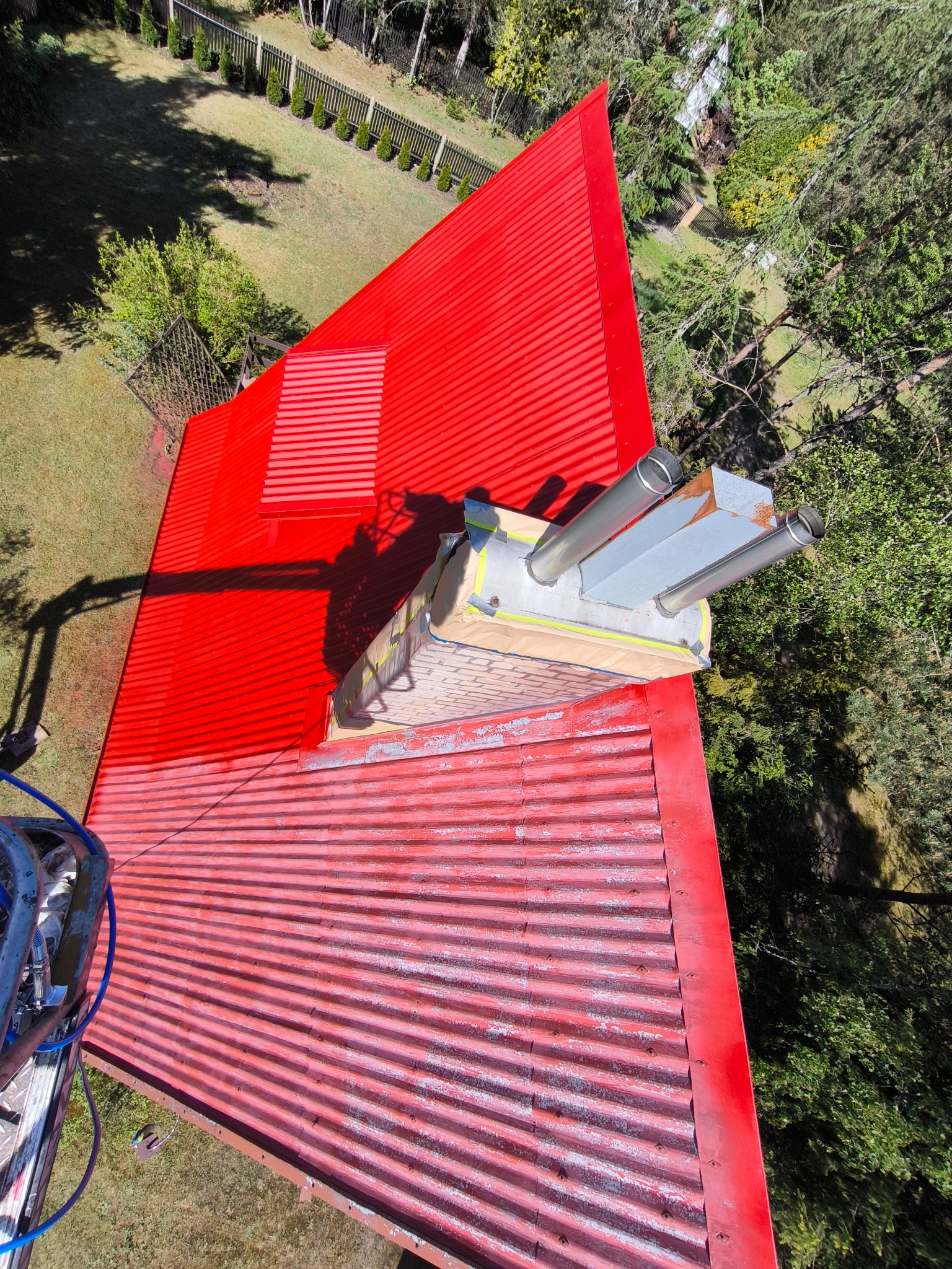 Czerwony, blaszany dach z kominem w trakcie renowacji, częściowo pomalowany, widoczne ślady starej farby i zabezpieczenia komina taśmą malarską, ujęcie z góry z widocznym otoczeniem drzew i trawnika.