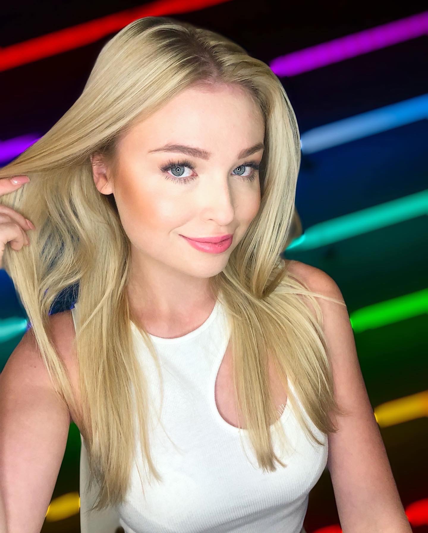 Portret uśmiechniętej blondynki z delikatnym różowym odcieniem ust, podkreślonymi rzęsami i rozświetloną cerą na tle tęczowych neonów.