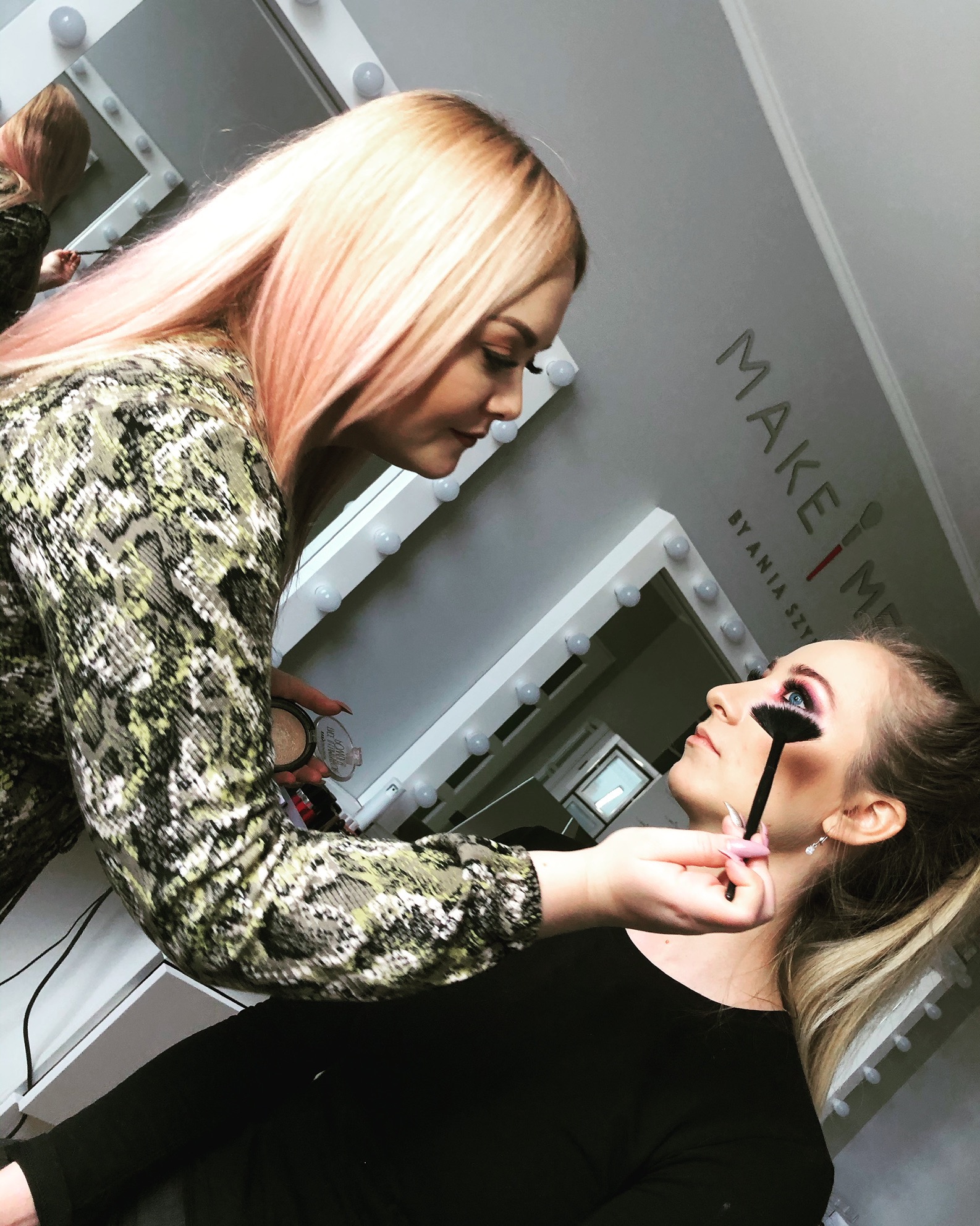 Profesjonalistka z blond włosami z różowymi pasemkami aplikuje pędzlem róż na policzki klientki w studio z lustrem do wizażu i napisem 'MAKE ME BY ANIA SZY'.