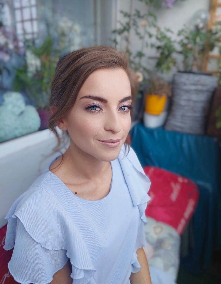 Portret kobiety o niebieskich oczach z delikatnym, świeżym upiększeniem, w błękitnej bluzce z falbanami, na tle balkonu z roślinami.