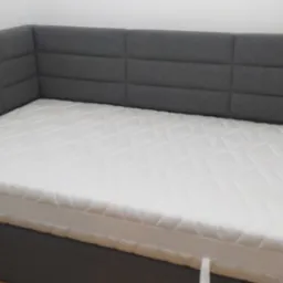 Szare, tapicerowane łóżko z zagłówkiem w kształcie paneli i białym materacem, widoczne w rogu pokoju.