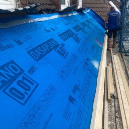 Usługi Budowlane Radosław Bersztolc - Montaż niebieskiej membrany dachowej Dasaplano 0.01 z widocznym pracownikiem na rusztowaniu i fragmentem starego dachu z dachówką.