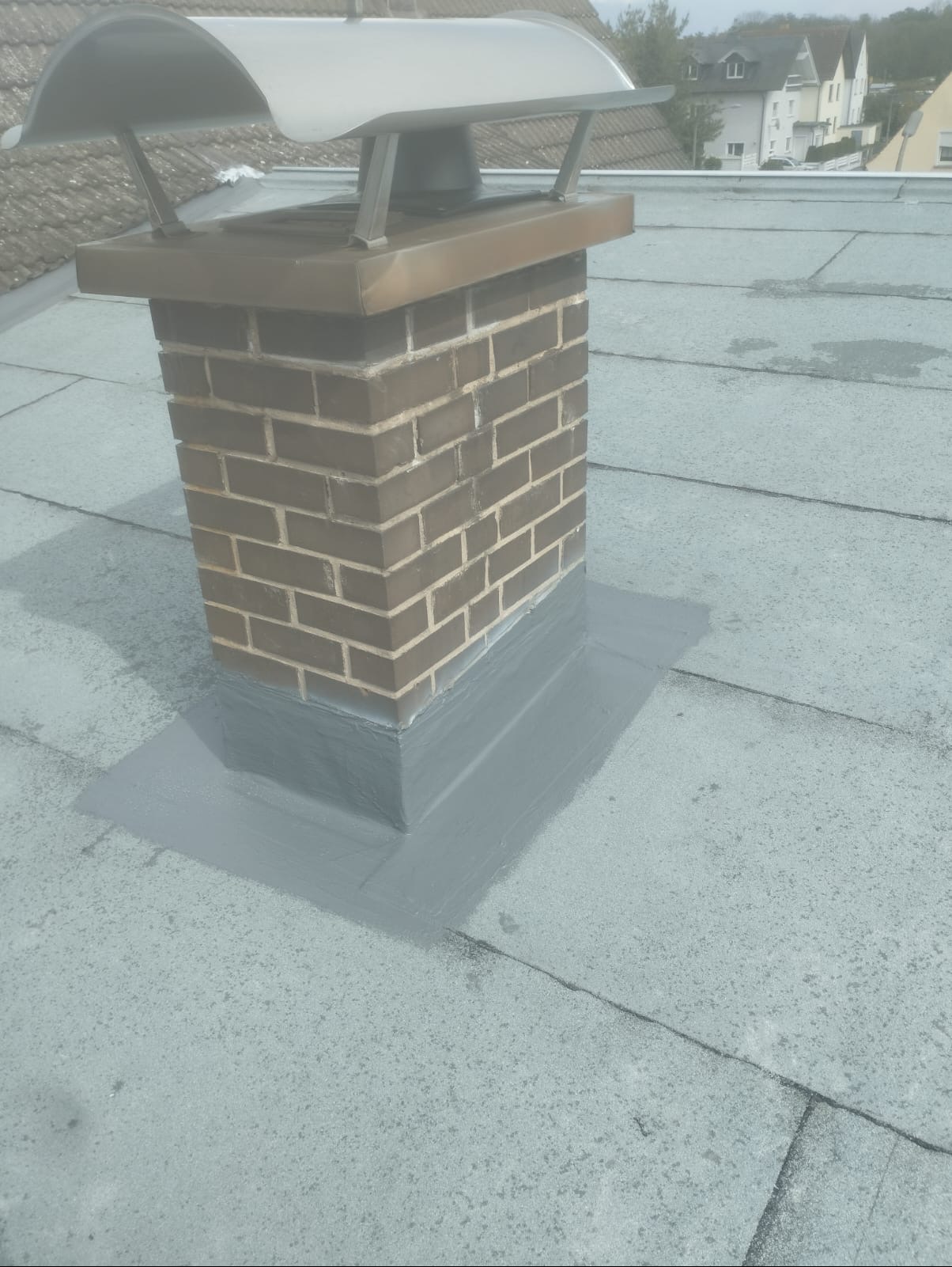 Kominek z cegły na płaskim dachu, uszczelniony szarą masą bitumiczną, z metalowym daszkiem zabezpieczającym przed deszczem.