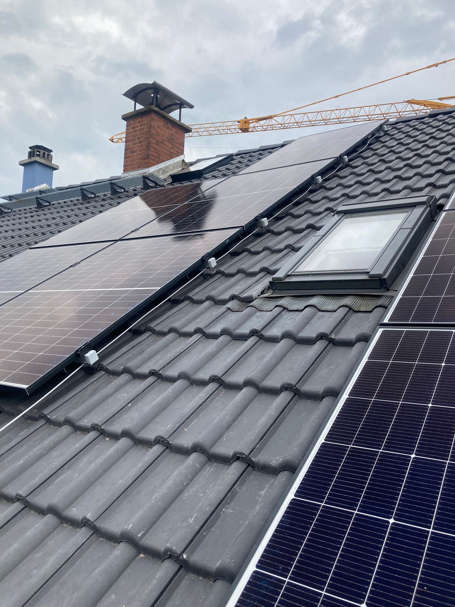 Czarne panele słoneczne zamontowane na ciemnoszarym dachu z dachówką ceramiczną, widoczny komin z cegły i żuraw budowlany w tle.