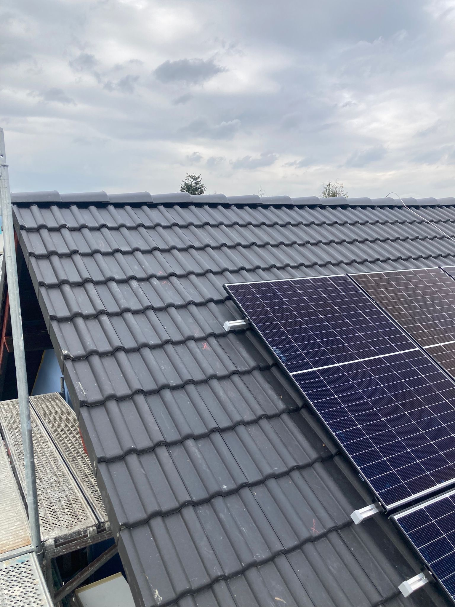 Ciemna dachówka ceramiczna z zamontowanymi panelami fotowoltaicznymi, widoczne rusztowanie i pochmurne niebo w tle.