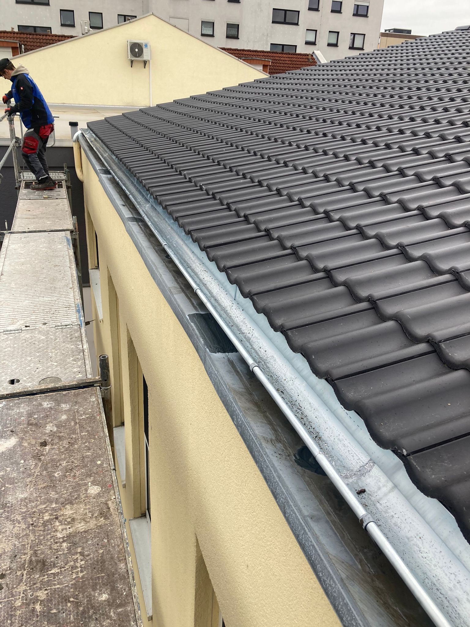 Pracownik na rusztowaniu montuje rynnę na krawędzi dachu pokrytego ciemną dachówką. Widoczny fragment elewacji budynku.