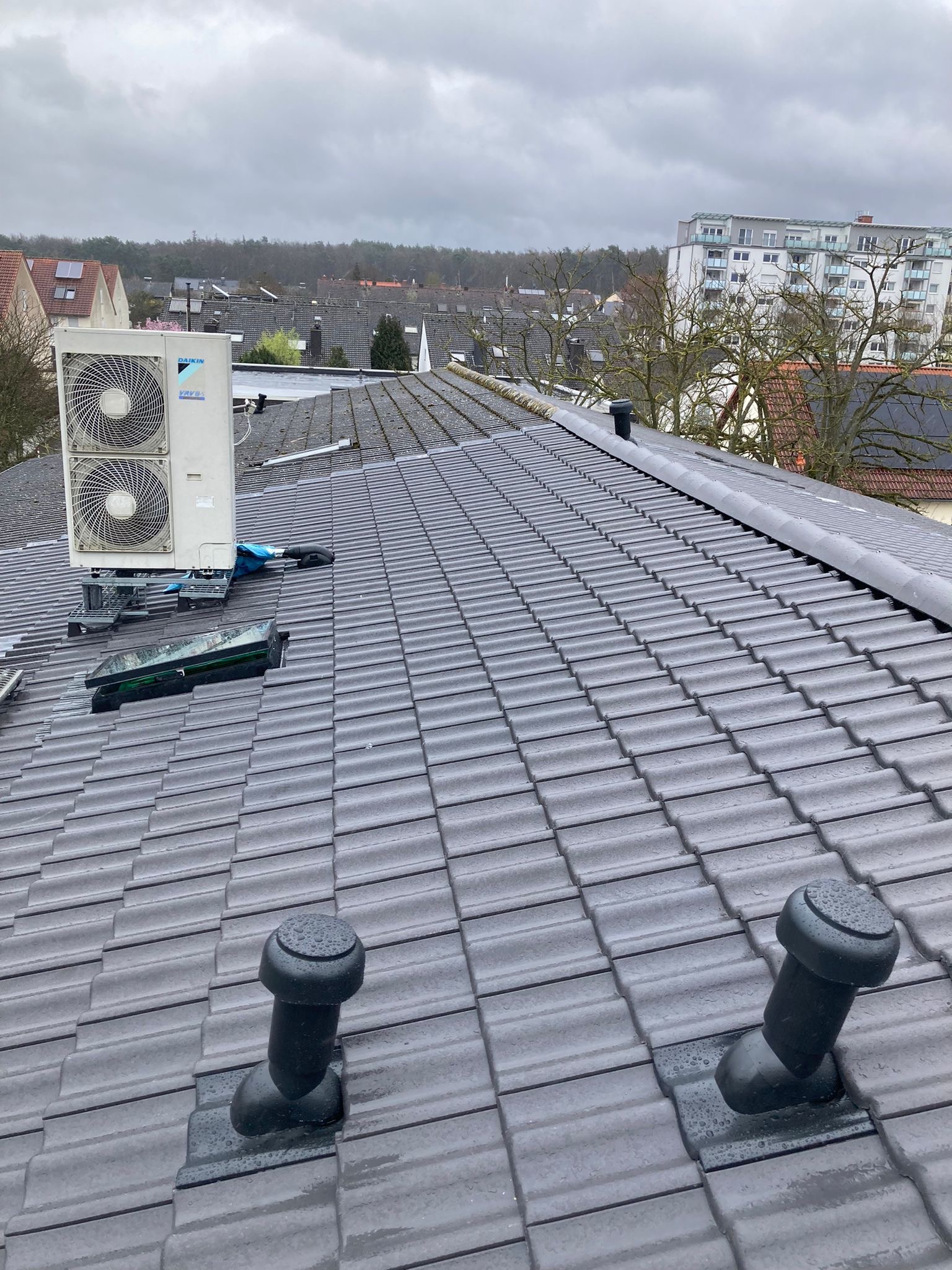 Szare dachówki ceramiczne na dachu z zamontowaną jednostką zewnętrzną klimatyzacji Daikin i wyłazem dachowym w pochmurny dzień.
