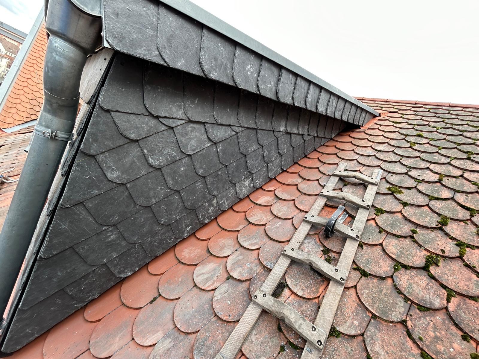 Fragment dachu z nową, łupkową obróbką blacharską przy kominie i starą, czerwoną dachówką ceramiczną pokrytą mchem, z drewnianą drabiną opartą o połać.